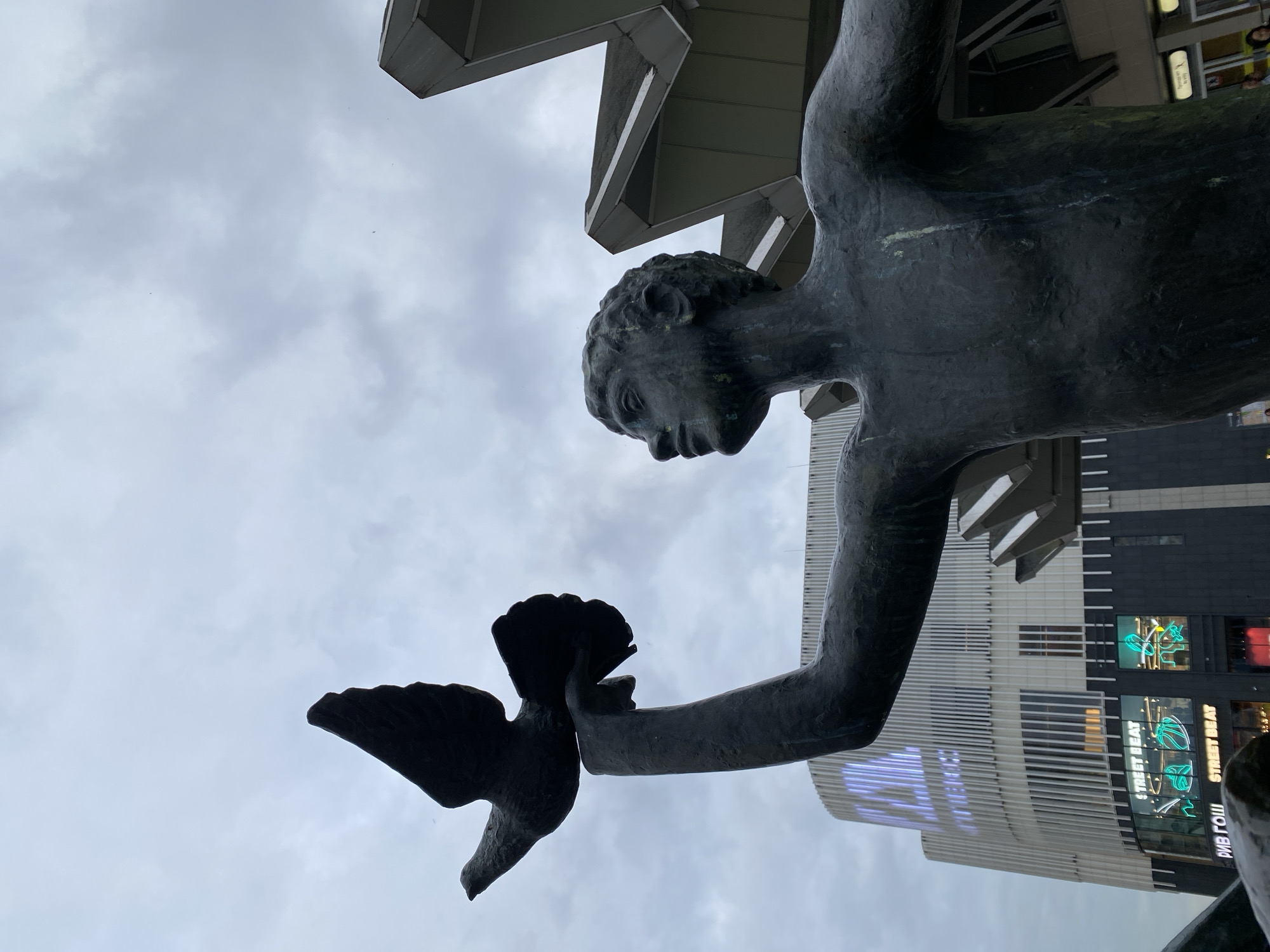 The image shows a statue of a man holding a bird, likely a dove, in his hand. The statue is bronze-colored and appears to be weathered. In the background, there is a modern building with horizontal lines and signage. The sky is overcast. The statue is the "Youth of Russia" sculpture by Zurab Tsereteli, located in front of the Stockmann building in Saint Petersburg, Russia.