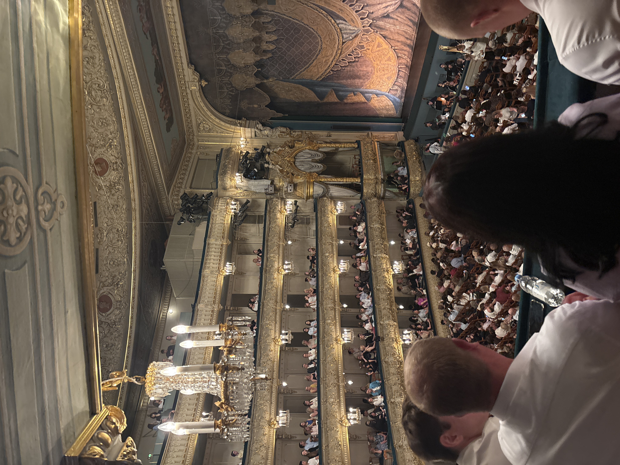 The image captures an interior view of the Bolshoi Theatre in Saint Petersburg, Russia, during a performance. The ornate architecture is evident, with multiple tiers of balconies filled with audience members. Chandeliers and decorative gilded elements adorn the ceiling and walls. The stage features a richly draped curtain. In the foreground, several individuals are visible from the back, attending the event. The audience is seated, with many people looking towards the stage or engaging with their phones. The overall scene depicts a cultural event in a grand historical venue.