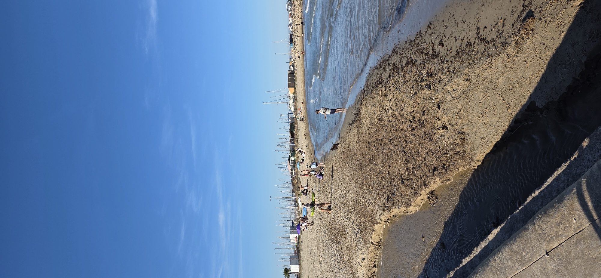 The image shows a beach in Canet d'en Berenguer, Spain. To the left is the open blue sky, transitioning to the beach, filled with beachgoers. There are numerous small boats lined up along the shore, suggesting the presence of a marina nearby. A person stands near the water's edge. A shadow is present at the bottom right corner. The beach is littered with seaweed.