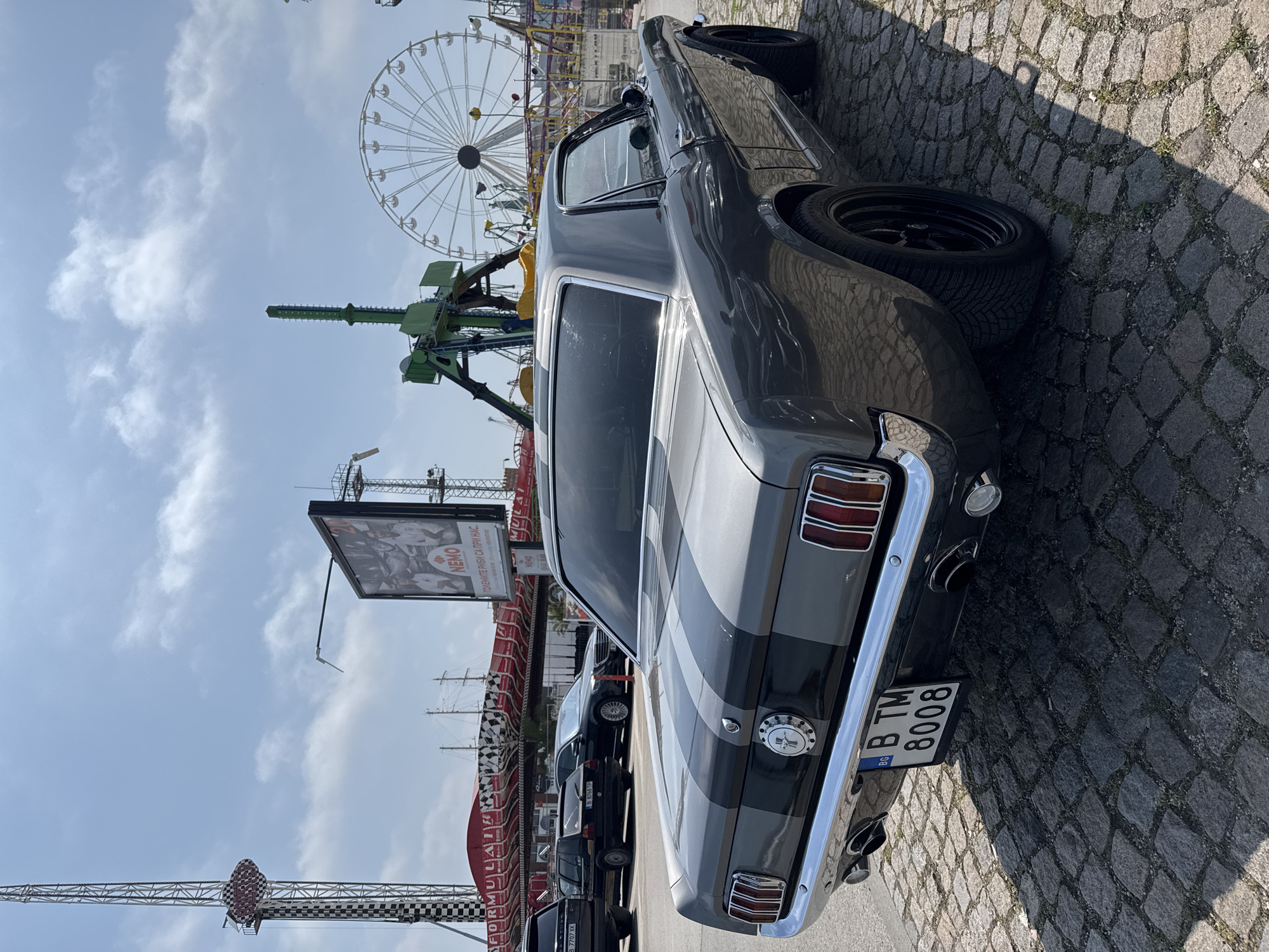 The image displays a classic dark grey car, likely a Ford Mustang, with two lighter grey racing stripes running longitudinally over its body. The car is viewed from its rear-passenger side, parked on a textured cobblestone surface. Its rear bumper, taillights, and a circular emblem are visible. The license plate reads "BG B TM 8008".
In the background, under a bright, partly cloudy blue sky, there is an amusement park or fairground. A prominent white Ferris wheel is visible on the left, along with a tall, green thrill ride, possibly a drop tower. Other structures and partial views of rides are also discernible. To the right, a large billboard features the word "NEMO" and Cyrillic text, including "ОГРАНИЧИТЕ РИБИ СА ПРИ НАС" (possibly "Limit the fish are with us" or "Catch the fish with us") and "СОЛЕНИТЕ РИБИ" (salty fish), suggesting a marine-themed attraction. Several other cars are parked behind the main vehicle. The time of day appears to be bright daylight, with shadows indicating the position of the sun. No people are visible in the scene. The image itself is rotated vertically.