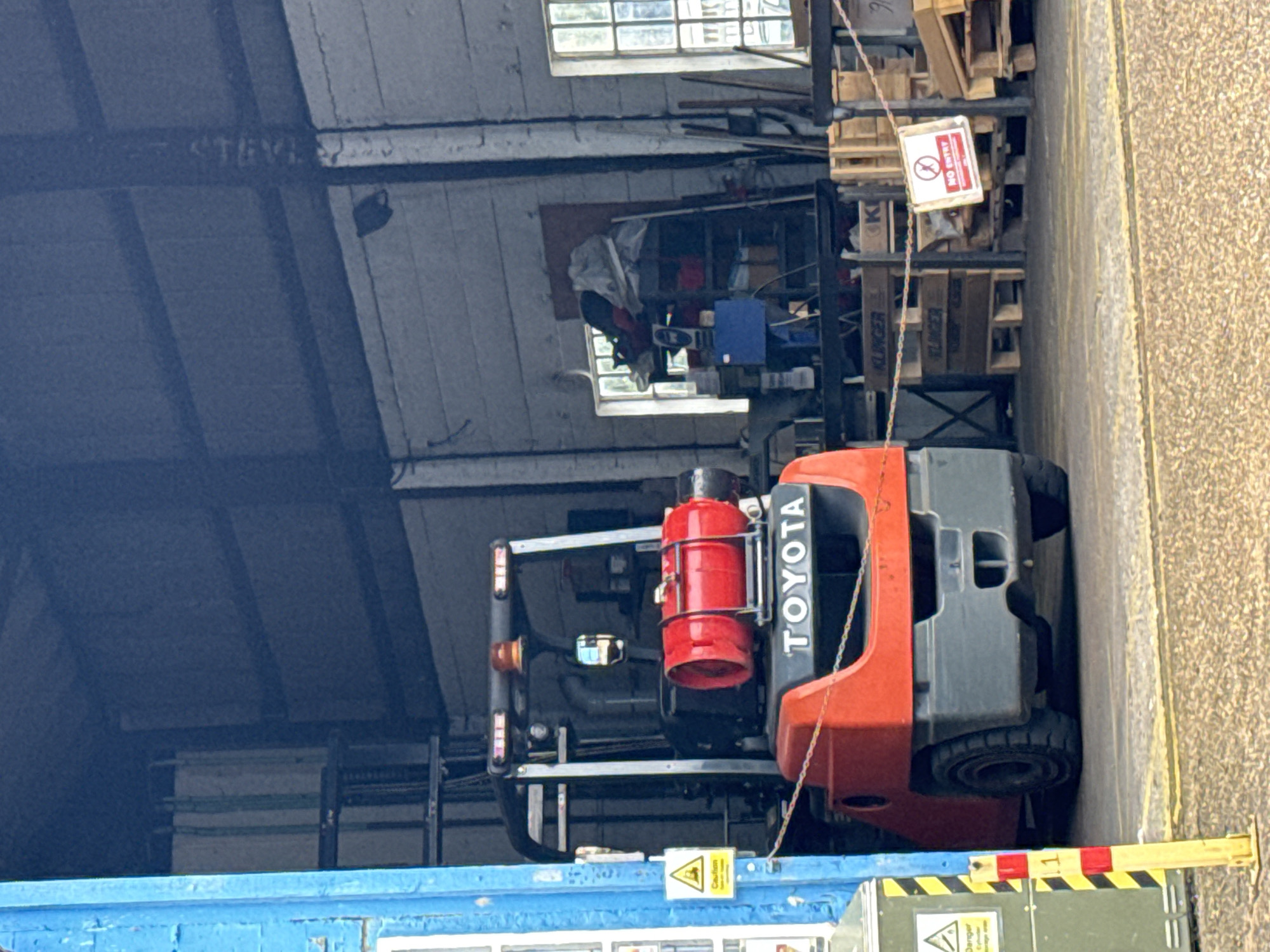 This image captures an industrial indoor setting, likely a warehouse or workshop, during daytime. The central subject is a red and grey Toyota forklift, which appears stationary and is positioned within or just outside the building. A red gas cylinder is prominently mounted on the rear of the forklift.
Daylight streams in through multiple windows visible in the background. Behind the forklift, various industrial items are stored, including stacks of wooden pallets, some of which bear the name "KLINGER". Miscellaneous objects like a blue box and a white protective covering are also visible on shelving. A white sign with a red border, hanging near the pallets, clearly displays "NO ENTRY" along with a prohibited pedestrian symbol. Faintly written on an overhead beam in the upper left background, the name "STEVE" can be seen.
In the immediate foreground, a blue painted structure, possibly a wall or a barrier, is present. Attached to this structure is a yellow triangular warning sign, partially readable as "Caution" and likely "Risk of injury". To the right of the forklift, a paved area, potentially an exterior access route, is visible.
The scene depicts a functional, industrial environment without any visible human activity. The overall impression is one of a working or storage area, consistent with the provided location context of Great Yarmouth, United Kingdom.