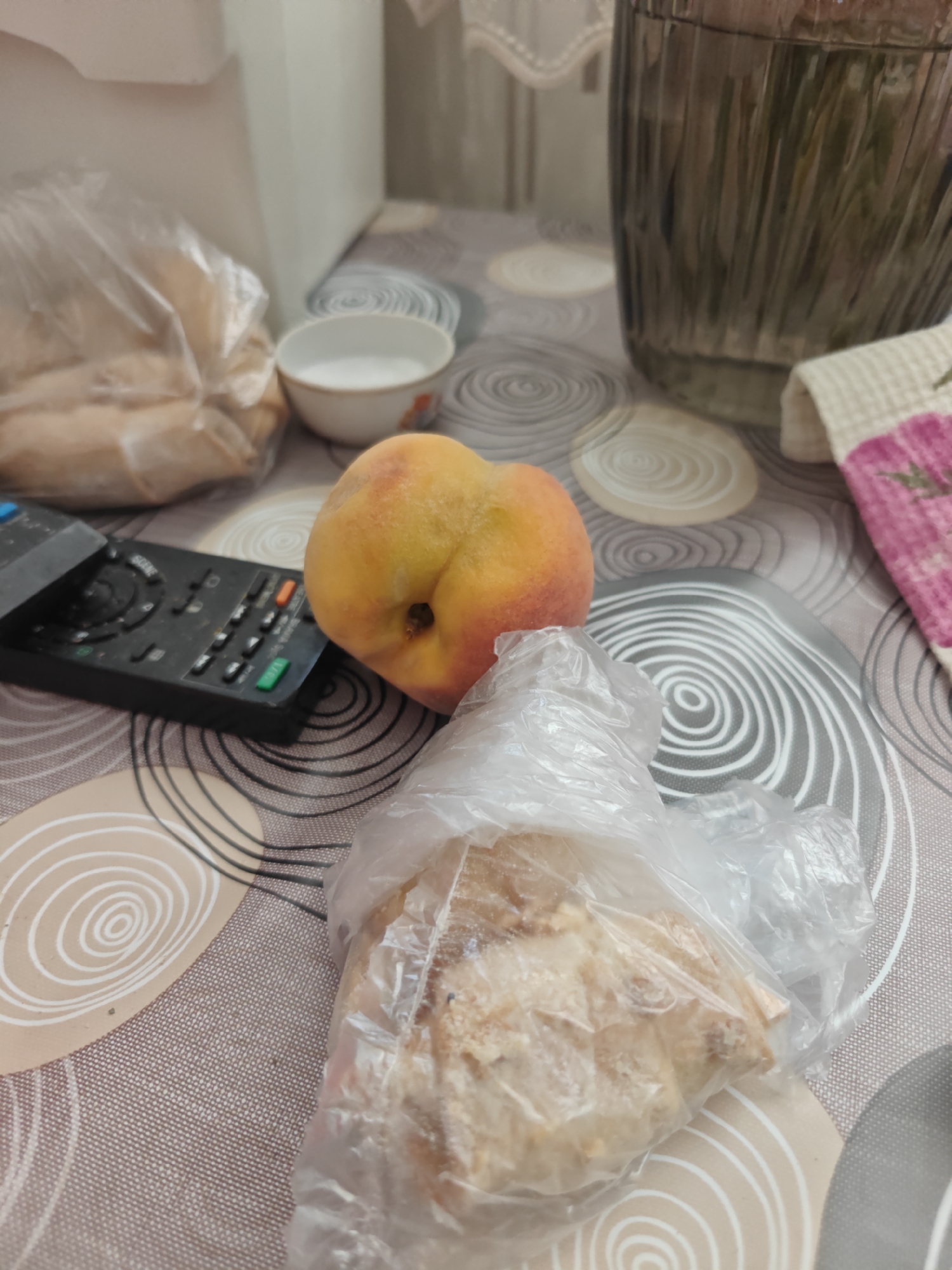 A close-up, indoor shot shows a peach, a remote control, a bowl of white substance, and a plastic-wrapped pastry on a patterned tablecloth. The peach is a vibrant yellow and red, with a visible indent. The remote is black with numerous buttons. The bowl appears to contain salt or sugar. The pastry is a light brown color and is partially visible through the plastic wrap. The tablecloth has a circular, swirling pattern in shades of beige and gray. In the background, a partially visible glass container and a white cloth with a pink and green pattern are present. The lighting is artificial and casts soft shadows. The scene is likely in a home environment in Krasnodar, Russia, given the general context.