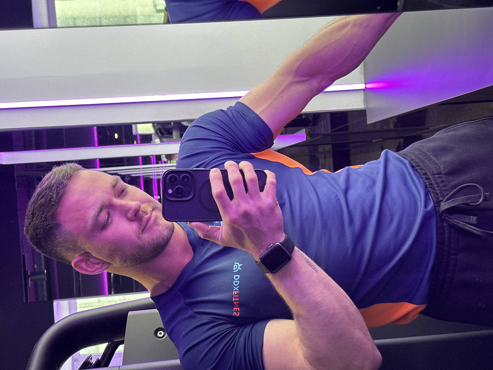 The image shows a man taking a selfie in a gym. The man is fair-skinned, with short, dark hair and a trimmed beard. He is wearing a blue and orange athletic shirt, dark athletic shorts, and a smartwatch on his left wrist. He is holding a black smartphone in his right hand to take the photo. The logo "DDXFITNESS" is visible on his shirt, reversed due to the mirror reflection. A tattoo is visible on his left forearm. The gym setting features workout equipment, a mirrored wall, and purple LED lighting. The location is presumed to be a DDXFitness gym, although its specific location in Perm, Russia is not conclusively discernable from the image.