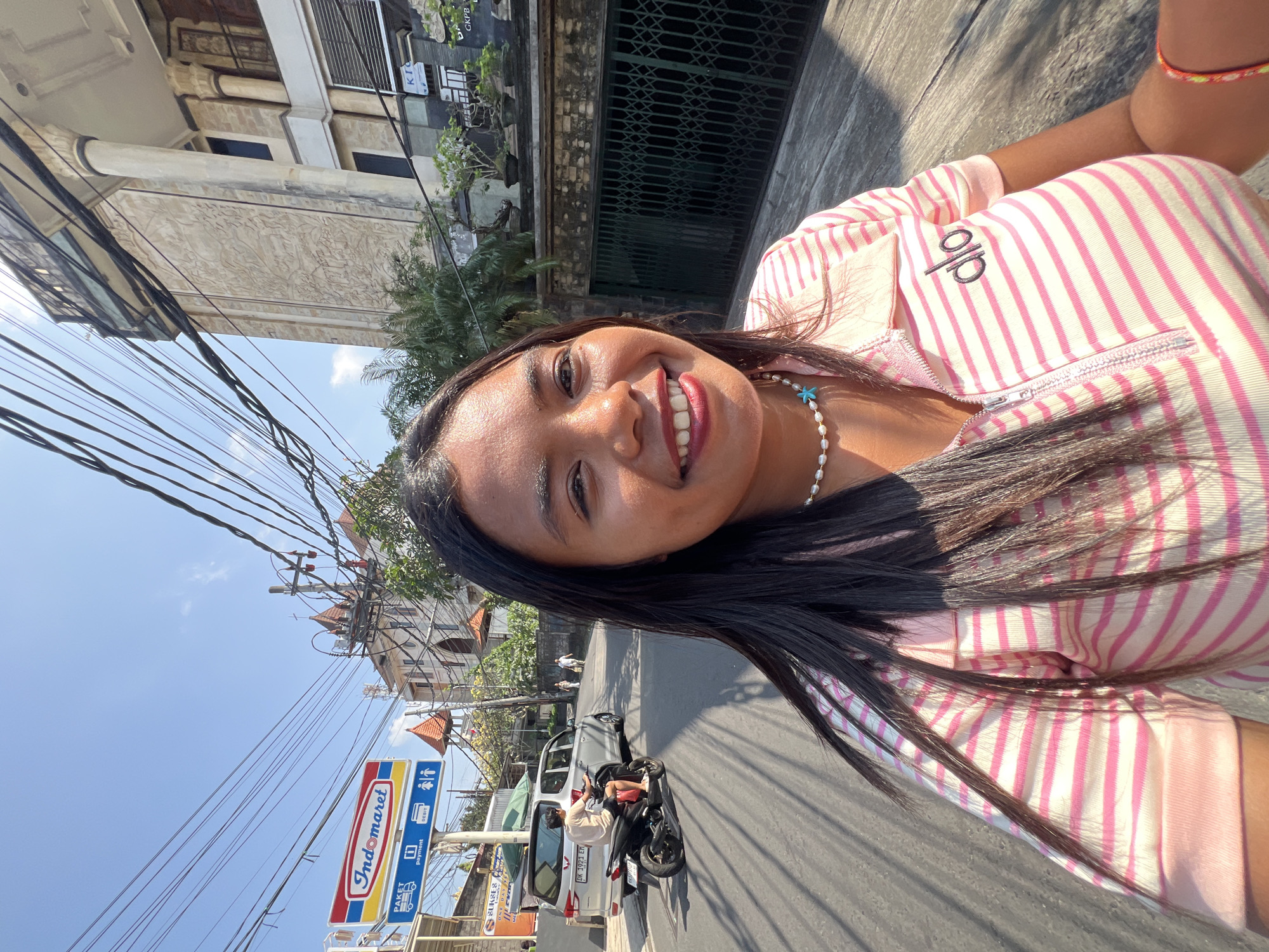 A woman with dark hair and a pearl necklace smiles into the camera, her face filling the right side of the frame. She is wearing a pink and white striped polo shirt with a zipper and a logo on the left chest. The background is a street scene in Kuta, Indonesia. To the left, an Indomaret convenience store sign is visible above a silver minivan. A person is riding a black scooter in front of the minivan. Buildings with intricate architectural details line the right side of the street, with power lines crisscrossing overhead. Sunlight casts shadows on the street, indicating it is a bright, sunny day.