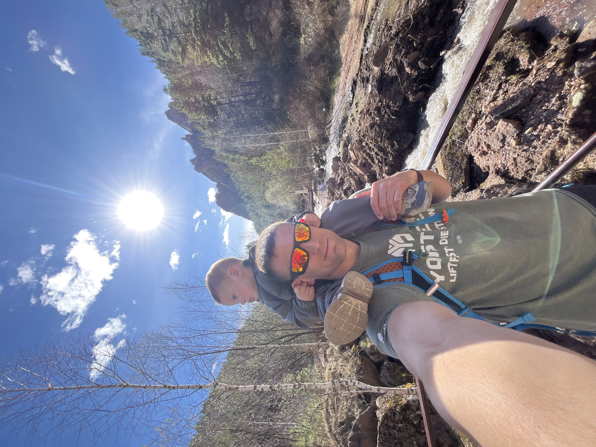 A man is carrying a young child on his shoulders. They are standing near a waterfall surrounded by rocky terrain and trees. The sky is blue with a bright sun. The man is wearing sunglasses, a green t-shirt, and has a backpack. The child is wearing a gray top. The photo is taken at an angle. Location is not explicitly identifiable as Krasnoyarsk, Russia.