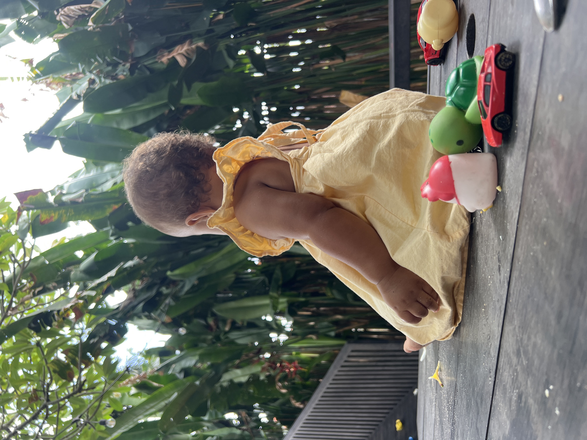 A young child, likely a toddler, is depicted from the side, wearing a yellow dress. The child is on a dark wooden surface, possibly a table or deck, surrounded by a lush, green, tropical environment. The background is filled with large, vibrant green leaves, suggesting a garden or outdoor space. Sunlight filters through the foliage, creating dappled light and bright highlights, indicating it is daytime, likely sunny.
Scattered on the wooden surface are several small toys, including a red toy car, a green spherical toy, and a pink and white object that resembles a cupcake or ice cream cone. There's also a yellow object, possibly a toy car accessory or a small toy figure. The child appears to be leaning forward, possibly reaching for or playing with these toys, though their expression is not visible. The overall scene evokes a sense of relaxed, outdoor play in a warm, tropical setting. The location context of North Kuta, Indonesia, aligns with the visual cues of abundant tropical foliage and outdoor living. There is no visible text in the image.