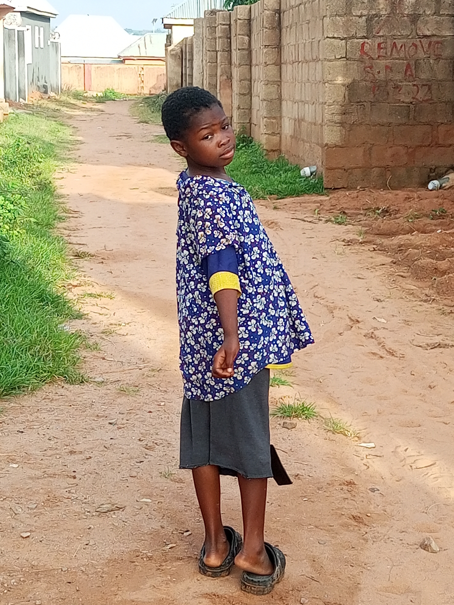 A young child, appearing to be under ten years of age, stands on a dirt path in what is identified as Tempure, Nigeria. The child is looking over their left shoulder towards the camera, with an inquisitive expression. They are wearing a blue and white floral print top with yellow accents on the sleeves and a dark grey skirt. The child is also wearing dark-colored slip-on sandals.
The environment is a dusty, unpaved road, with sparse green vegetation lining the left side of the path. To the right of the child, a rough, brick wall is visible, bearing red graffiti. The wall appears to be part of a building structure. In the background, more buildings with corrugated metal roofs can be seen, suggesting a residential area. The sky is light and overcast. The overall scene depicts a child in a modest, likely rural or semi-urban, setting in Nigeria.