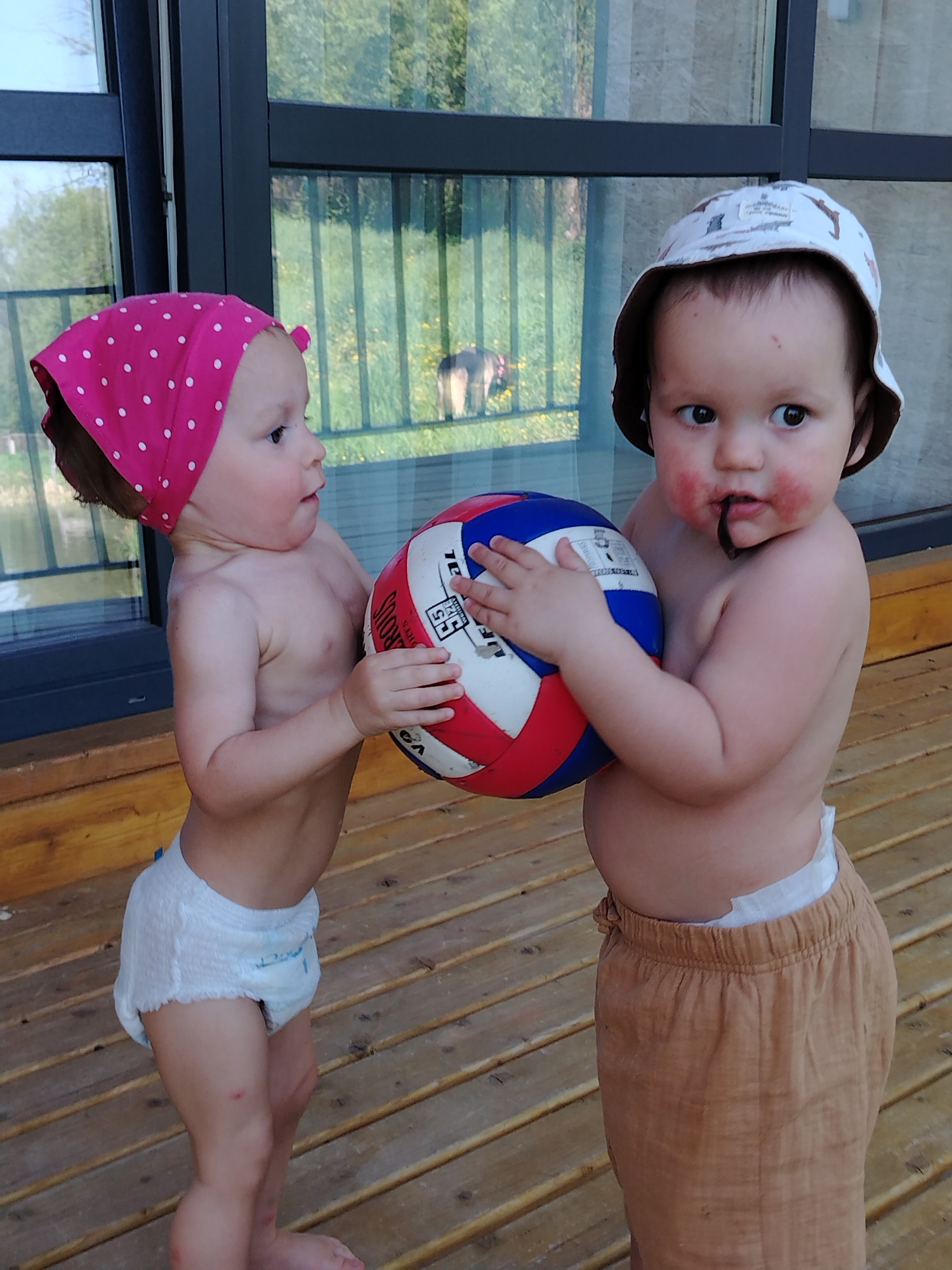 Two toddlers stand on a wooden deck, likely outdoors. The child on the left wears a pink polka-dot headscarf and a diaper, reaching for a red, white, and blue volleyball. The child on the right wears a light-colored hat and brown shorts over a diaper, holding the same volleyball. A dark object is in the mouth of the child on the right. In the background, a building with glass windows reflects an outdoor scene with grass and an animal. The location is Malyi Bukor, Russia.