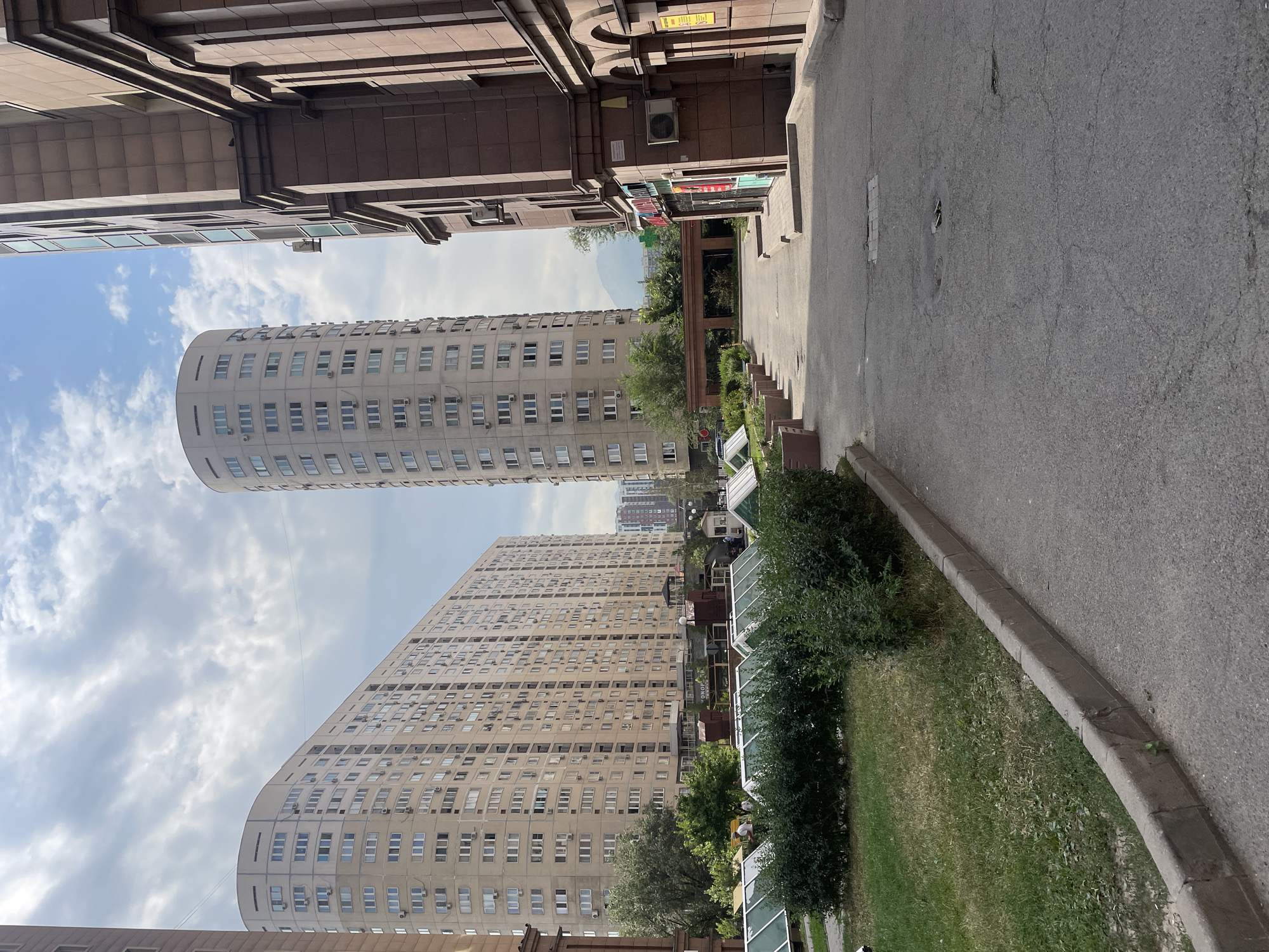 The image depicts a street scene in Almaty, Kazakhstan, likely in a residential or commercial area characterized by modern architecture. In the foreground, a paved walkway curves to the right, bordered by a grass strip and hedges. Several low, rectangular structures with glass roofs are visible in the grass area, possibly representing greenhouses or skylights for underground spaces.
Dominating the mid-ground and background are several large, multi-story buildings. On the left, a long, curved building with numerous windows and air conditioning units extends across the frame. To its right, a cylindrical tower building, also featuring many windows and visible air conditioning units, stands tall. In the far background, another residential building is visible, as is a mountain range under a cloudy sky.
On the right side of the image, a brown, stone-clad building with arched entrances and windows occupies the edge of the frame. A street sign and storefront signage are partially visible on this building. The overall impression is one of an urban environment with a mix of residential and commercial structures, situated in a city with mountainous surroundings. The lighting suggests it is daytime, with scattered clouds in the sky. There are no people clearly visible in action, though a few figures may be present in the distance.