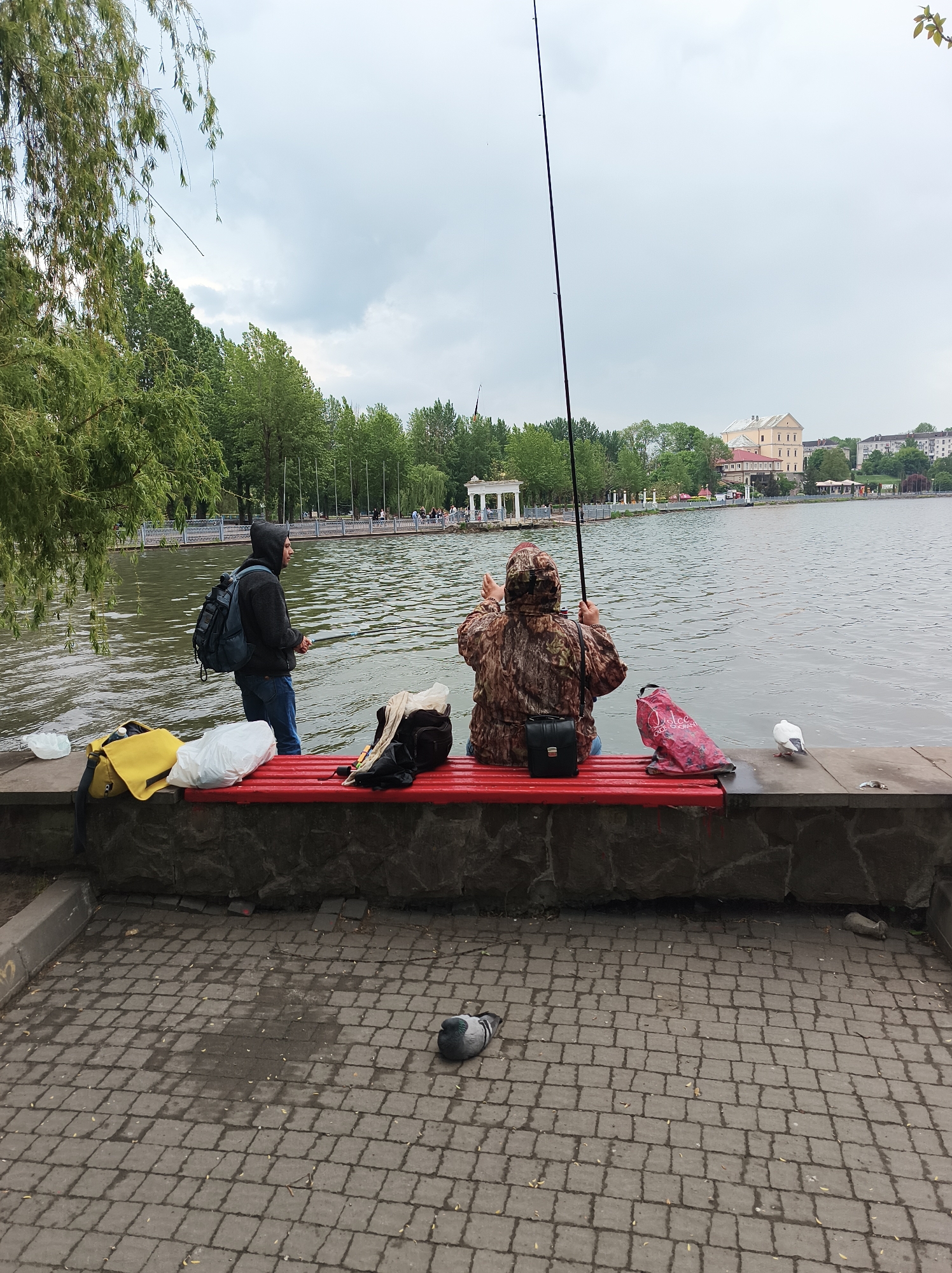 Two people are fishing from a red bench along a waterfront. One person stands wearing a black hooded sweatshirt and blue jeans, facing the water with a fishing rod. A backpack rests on their back. To their right, another person sits on the bench wearing a camouflage jacket and jeans. They hold a fishing rod. A small black bag and a patterned red bag are on the bench next to them. Various bags and other items are scattered on the bench and the ground nearby. A white pigeon stands on the edge of the water. A gray pigeon sits on the brick pavement in the foreground. The body of water is a lake with a light-colored gazebo on the opposite shore. Buildings and trees are visible in the background. The scene is overcast. This is the Ternopil Lake in Ternopil, Ukraine.