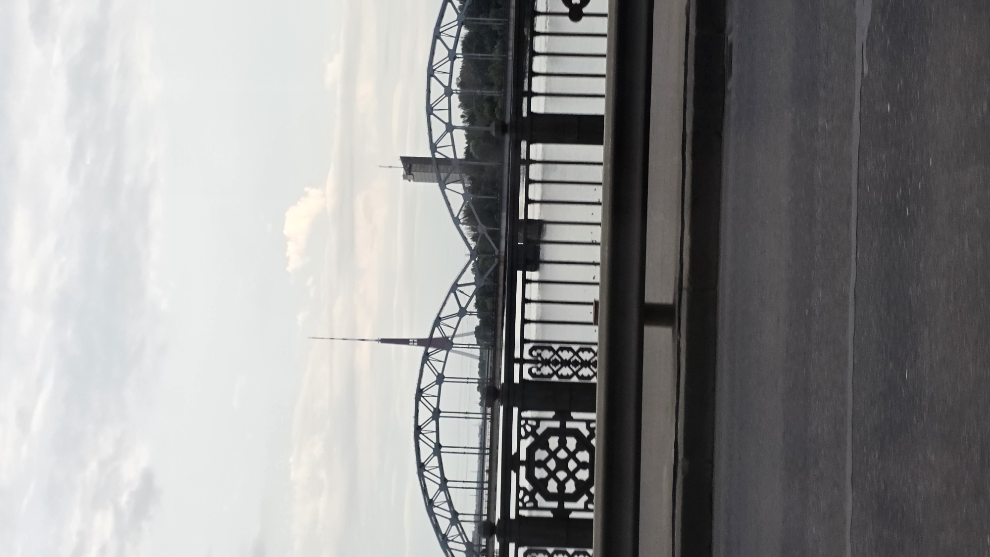 The image depicts the Riga TV Tower and a modern building as seen from the Riga Central Market bridge in Riga, Latvia. The bridge features ornate black railings with decorative patterns and a gray concrete barrier. The Riga Central Market is a large, domed building visible behind the bridge. The Riga TV Tower, a tall, slender structure with a red top, stands prominently in the background, along with a more recent, rectangular building. The scene is set against a cloudy sky and a body of water, likely the Daugava River. The foreground shows a road, suggesting the image was taken from a moving vehicle.