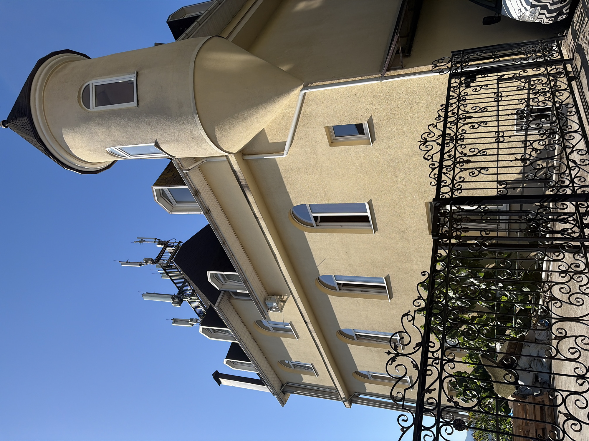 A low-angle, full shot shows the side of a light yellow, multi-story building with a dark grey roof, set against a clear blue sky. The building features a rounded turret on the right, topped with a dark grey conical roof. Several windows with white frames are visible on the building's facade. On the roof, a metal structure supports multiple cell phone antennas. In the foreground, a black wrought iron gate with ornate scrollwork spans across the bottom of the frame. Behind the gate, greenery is visible along with some stacked wooden pallets and large white bags. The scene appears to be in a coastal city, possibly Sochi, Russia, given the architecture and the presence of the sea in the distance.