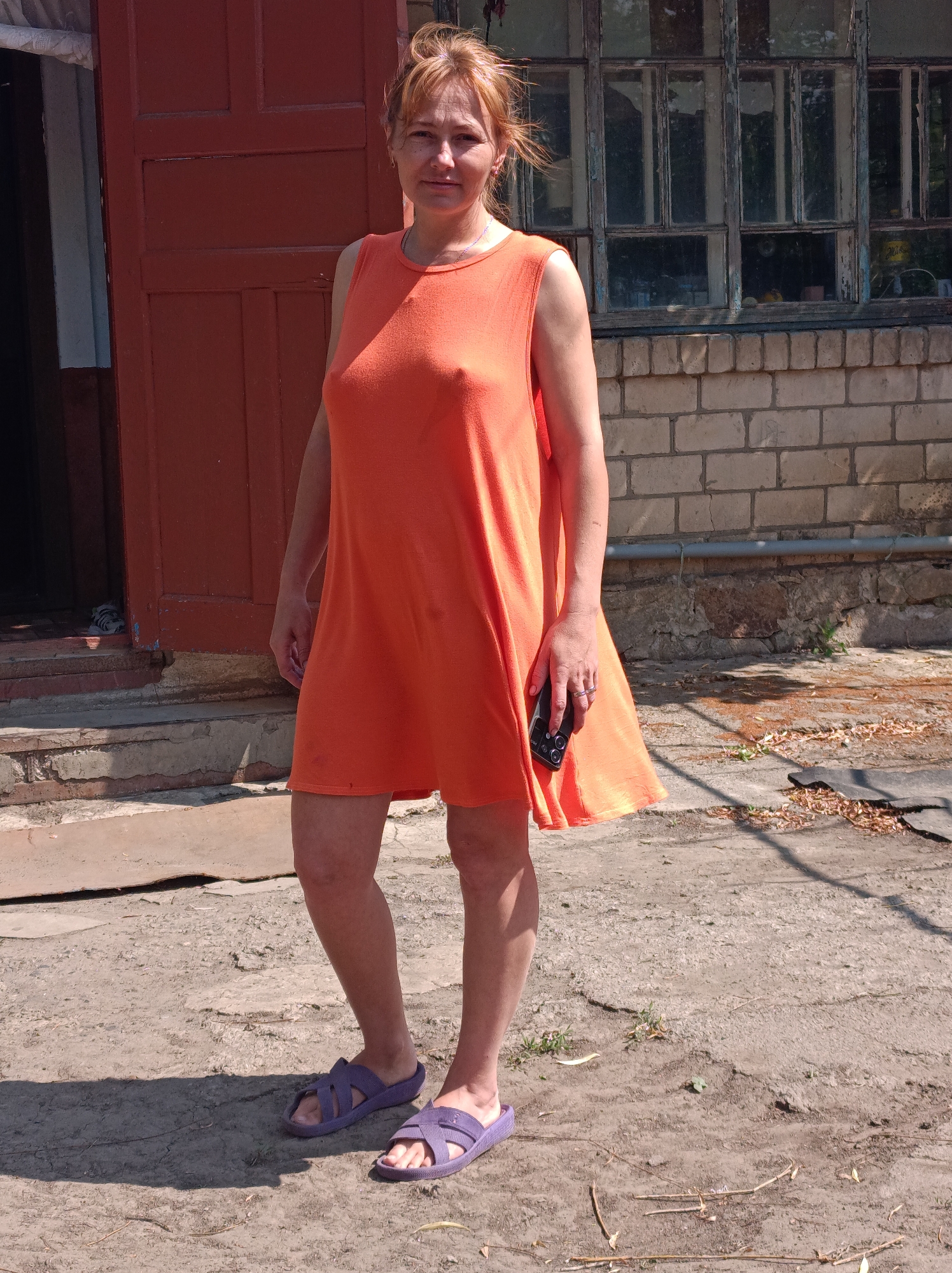 A middle-aged woman with blonde hair pulled back stands in front of a weathered brick building with a large, multi-paned window. She is wearing a sleeveless orange dress and purple sandals. Her left hand holds a black mobile phone. The ground is a mixture of dirt and cracked concrete, with scattered dry leaves. The background suggests an outdoor setting, possibly a yard or alley, with an orange door visible to the left. The overall scene appears to be in an urban environment in Saksahan, Ukraine.