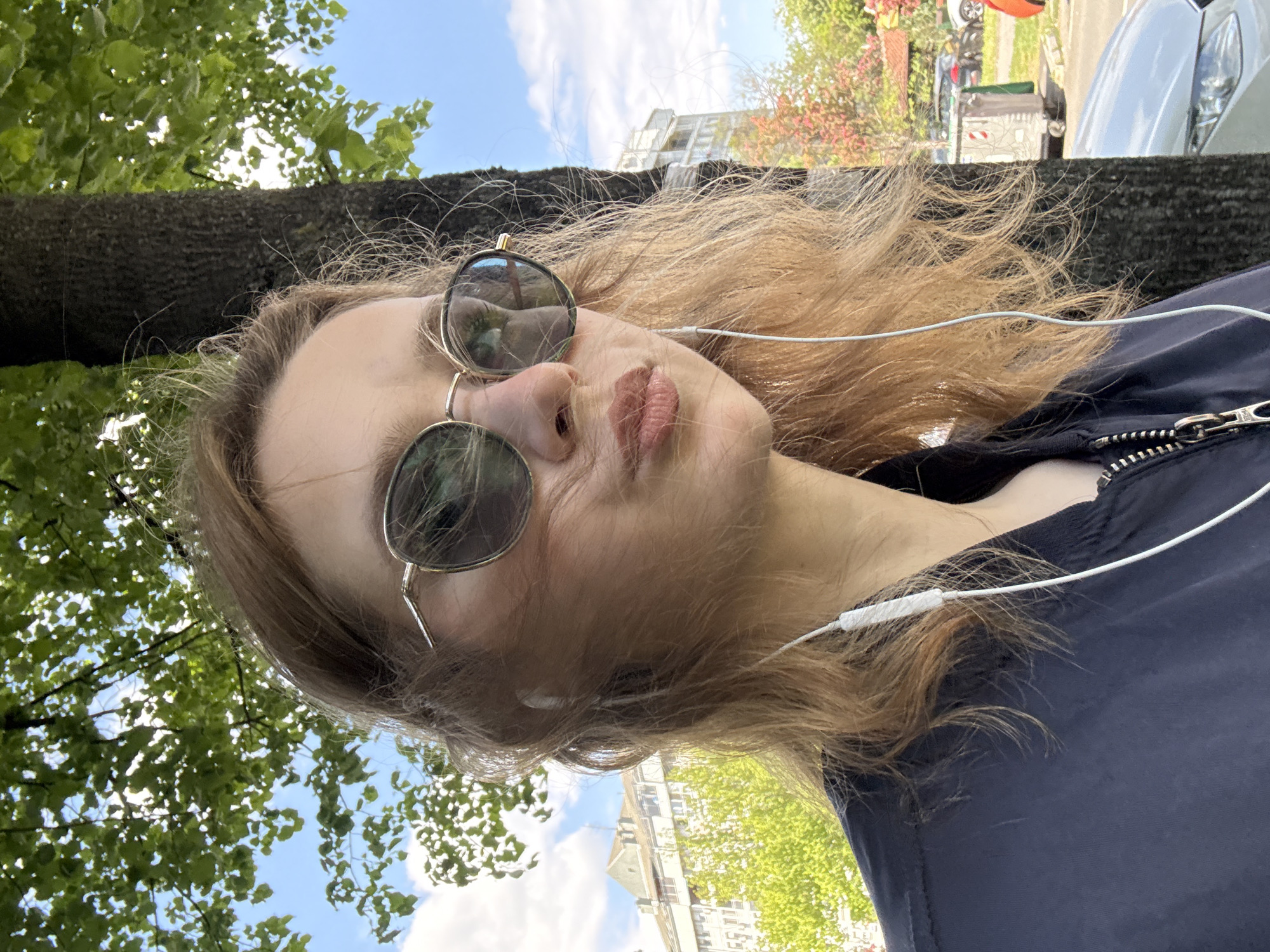 A young fair-skinned woman with light brown hair is wearing sunglasses and earphones while outside. The photograph appears to be taken at a low angle looking upward toward the woman's face. Behind her is a large tree with green leaves and a building in the background. There are also cars parked along the street, and the sky is visible. The woman is wearing a dark blue jacket with a zipper. Based on the architectural style and street features, the image may have been taken in the city of Belgrade, Serbia.