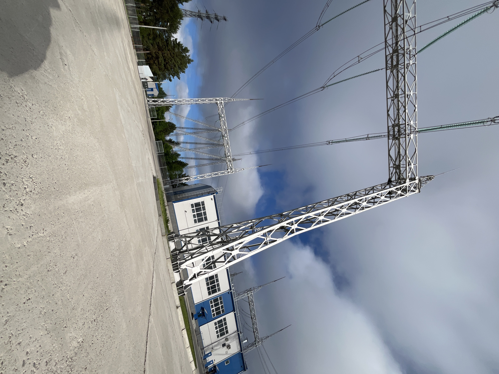 The image depicts an outdoor view of an electrical substation in Chaikovsky, Russia. Dominating the foreground is a vast expanse of light gray concrete ground, angled upwards towards the right. A large, white metal power line pylon, characterized by its lattice structure, extends diagonally across the frame, originating from the lower left and stretching towards the upper right. This pylon supports thick, dark electrical cables that run horizontally.
Behind the pylon, a modern, rectangular building with a white facade and a blue roof is visible. It features numerous windows, some with visible blinds or shades. Adjacent to this building, another, smaller white structure is present, with a single visible door.
In the background, more electrical infrastructure can be observed, including additional pylons and a forest of tall, green pine trees. The sky above is a mix of dramatic, dark gray clouds and patches of bright blue sky, suggesting changeable weather conditions. A distant view of another, larger electrical tower is also present to the left. The overall scene conveys an industrial and functional environment.