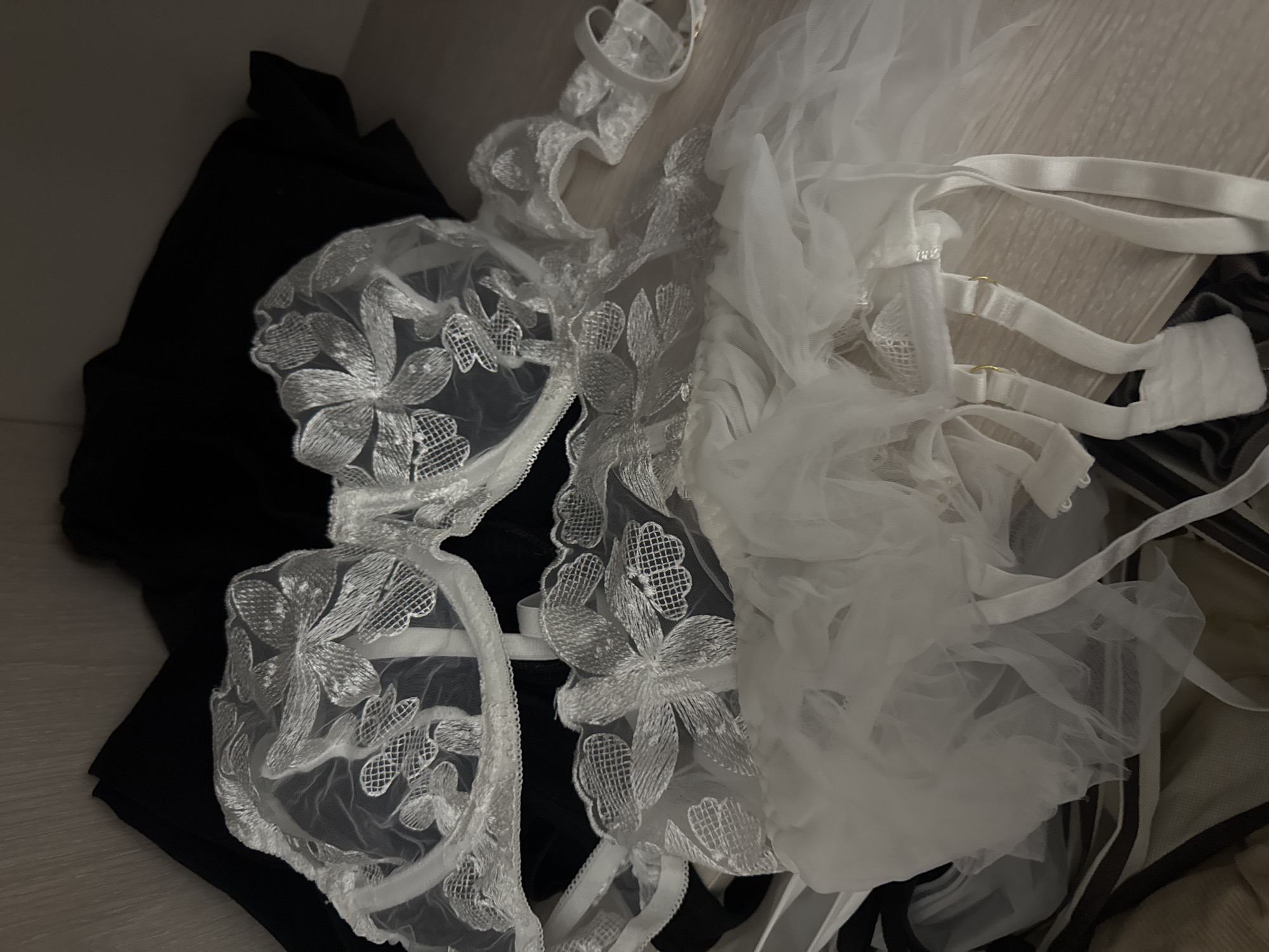 A white lace bra with floral embroidery is laid on top of a black garment, likely a dress or skirt. A white tulle garment with straps is also visible, partially obscuring the bra. The scene appears to be inside a closet or drawer, with wooden shelves or a surface visible beneath the clothing. There is no indication of the location in Saratov, Russia, or any people present.