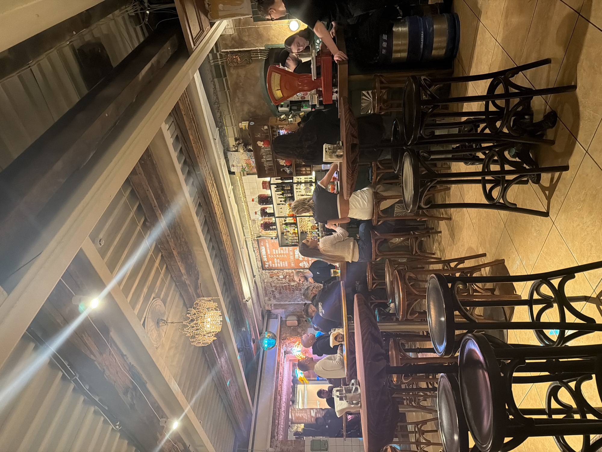 The image depicts the interior of a bar or pub in Moscow, Russia, likely during the evening. The focus is on the bar area where several patrons are seated on high stools. They are engaging in activities such as drinking and talking. Two bartenders are visible behind the bar. The establishment appears to be a mix of modern and rustic with exposed wooden beams on the ceiling, a chandelier, exposed brick walls and neon lighting, adding to the atmosphere. The floor is tiled. Bar stools of a classic bentwood design are a prominent element. A variety of bottles are displayed behind the bar.