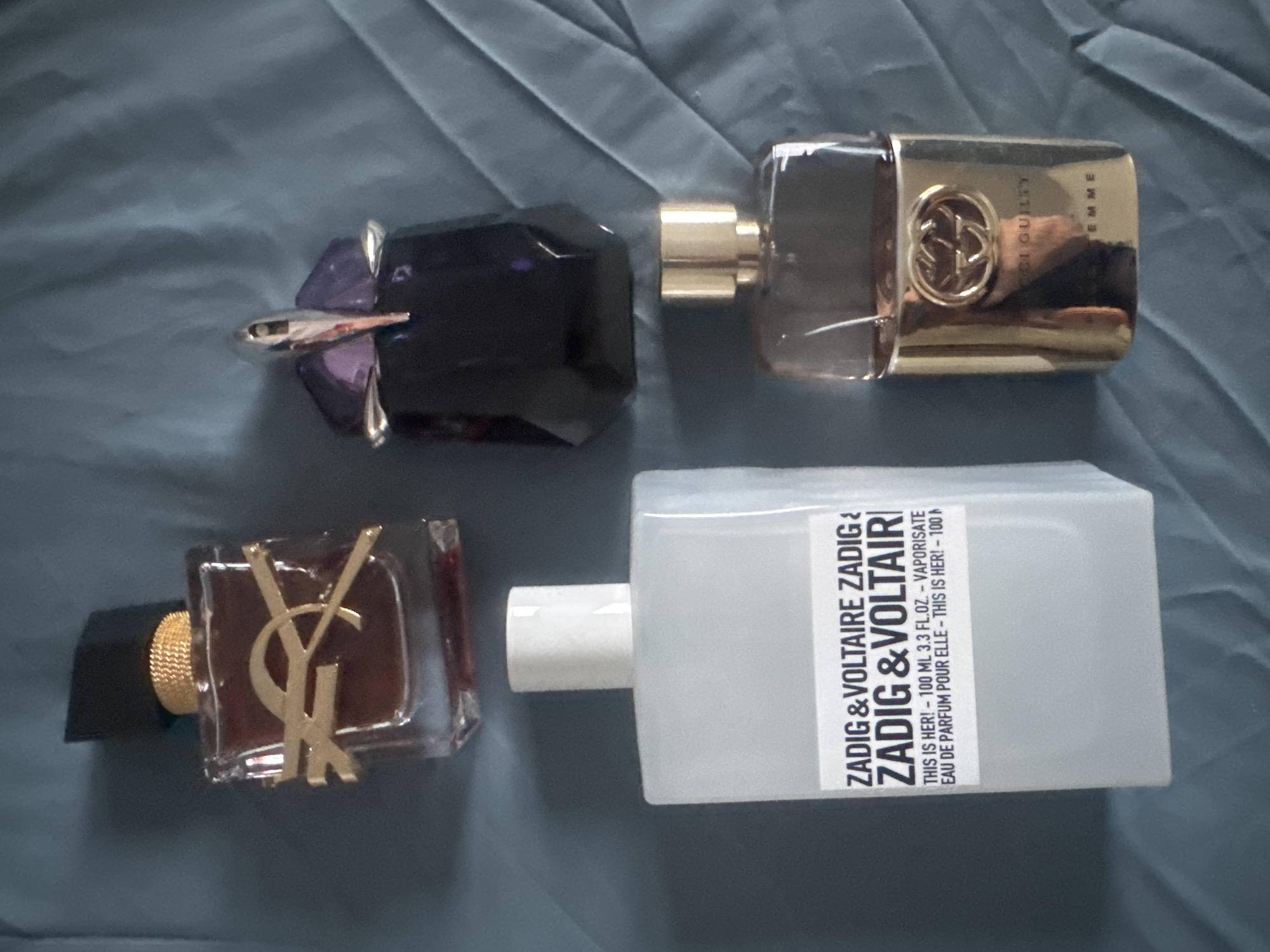 Four perfume bottles are arranged on a dark blue, textured surface. In the top left, a rectangular glass bottle with a gold accent and a black cap contains a reddish-brown liquid. To its right, a dark purple bottle with a metallic silver and red atomizer top is partially visible. In the bottom left, a tall, opaque white rectangular bottle has a white cap and a label with black text that reads "ZADIG & VOLTAIRE." To its right, a gold-colored glass bottle with a metallic gold cap has the Gucci logo embossed on its front, and text that appears to read "GUCCI GUILTY" and "FEMME."