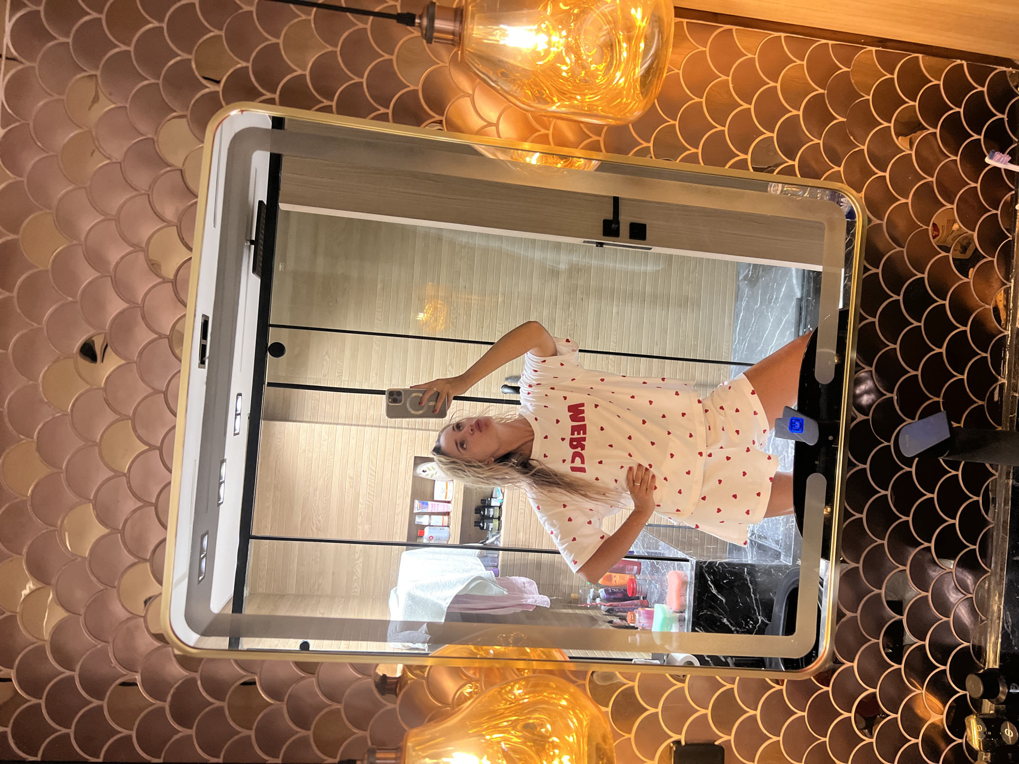 The image shows a woman taking a selfie in a bathroom. She is wearing pajamas with a heart pattern and the word "MERCI" printed on the top. The bathroom has a vanity with a mirror, sink, and various toiletries. The walls are decorated with fish-scale tiles in shades of brown and gold. There are two pendant lights hanging from the ceiling, emitting a warm, orange glow. The scene appears to be from a hotel bathroom in Sharm El Sheikh, Egypt, as indicated by the luxurious decor and the presence of items commonly found in a resort setting.