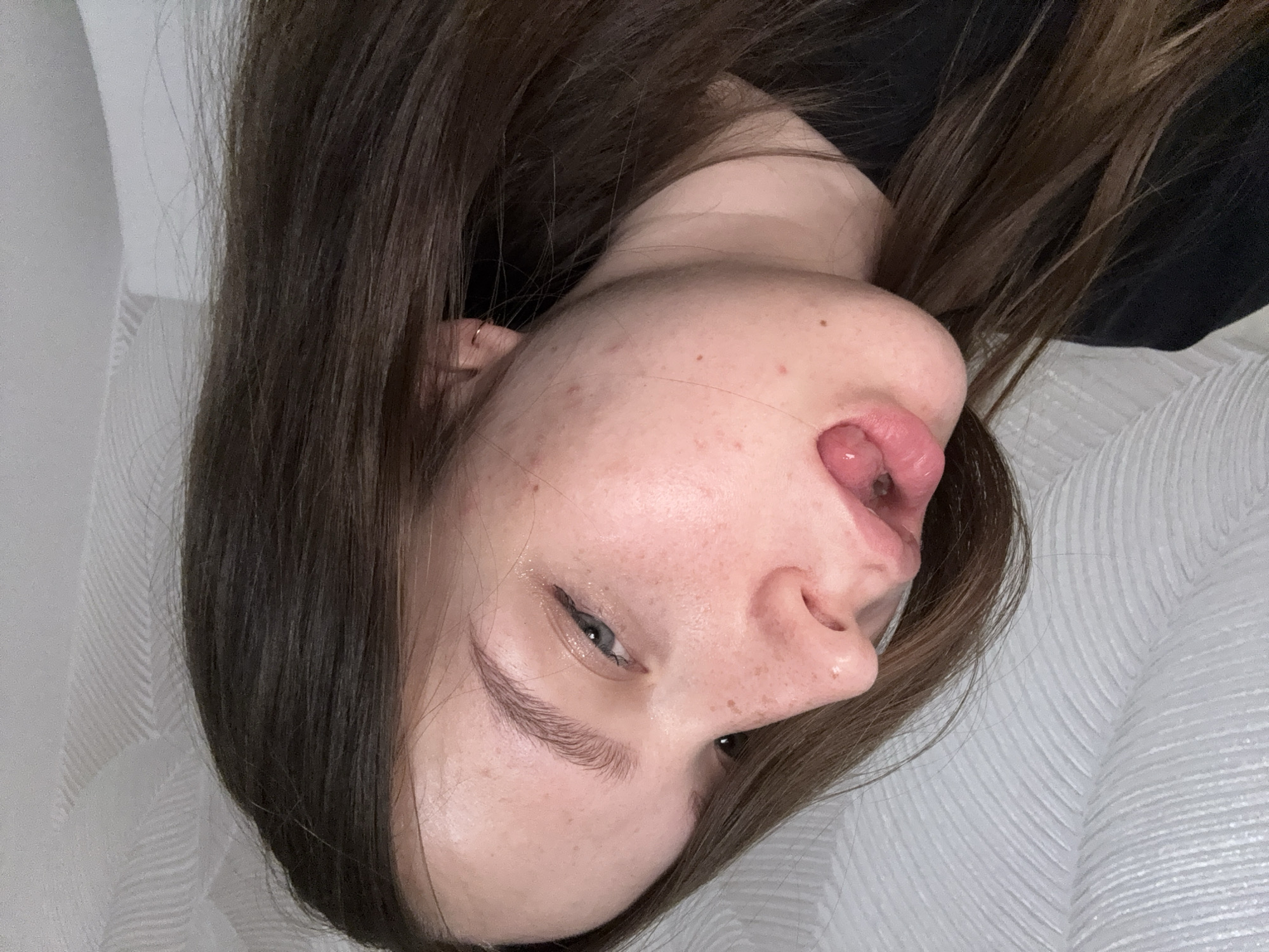 The media file presents a close-up portrait of a young person, likely female, captured from an overhead or slightly angled perspective. The individual is lying down, with their head tilted, looking directly towards the camera. They have long, straight, dark brown hair that falls around their face and extends across much of the frame.
The person's face shows several notable details. Their left eye (viewer's right) is open, revealing a light-colored iris, and features a subtle black winged eyeliner. Their eyebrows are dark and well-groomed. The most prominent feature is their mouth, with the tongue partially extended and curled upwards, touching the philtrum area just below the nose. The skin on their face appears natural, displaying visible freckles and a few minor blemishes. There is a slight, natural sheen on the skin, particularly on the forehead and around the eye. A small, delicate gold hoop earring is visible in their right ear.
The individual is wearing a dark-colored top, barely visible around the neckline. The background consists of a light, possibly white or off-white, textured surface, which could be bedding, a pillow, or a curtain, featuring a subtle repetitive wave or herringbone pattern. The overall scene suggests a casual, indoor setting, possibly in Chaikovsky, Russia, where the person is making a playful or expressive face. There is no visible text in the image.
