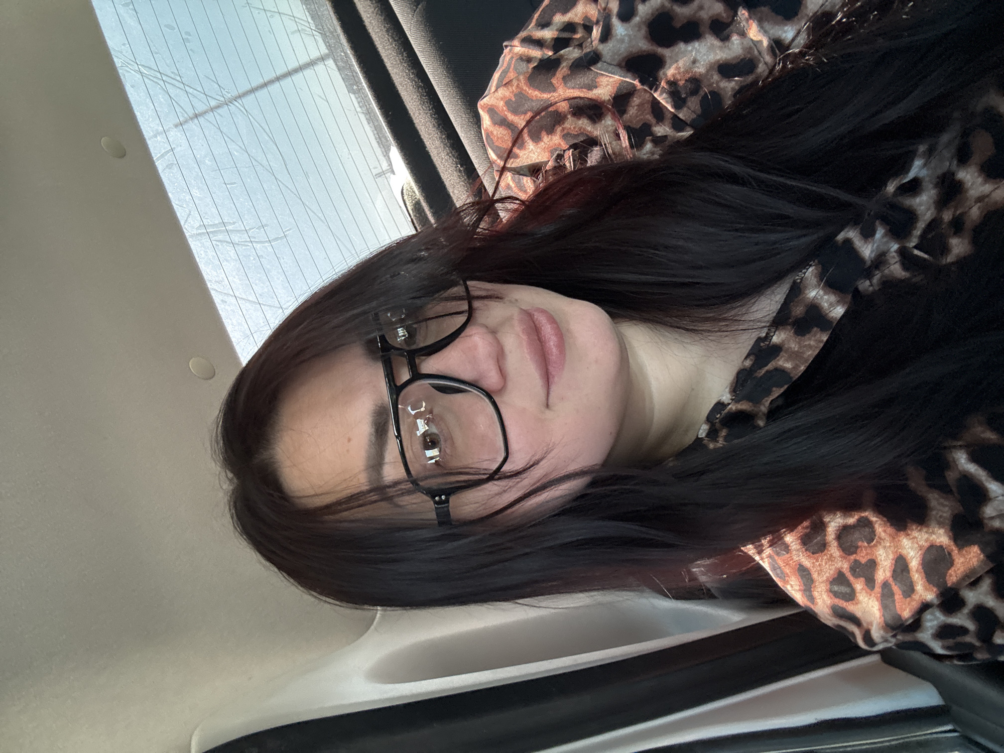 A woman with dark hair and glasses is in a car, wearing a leopard print top. The car window is visible in the background. The location is Unknown, Unknown.