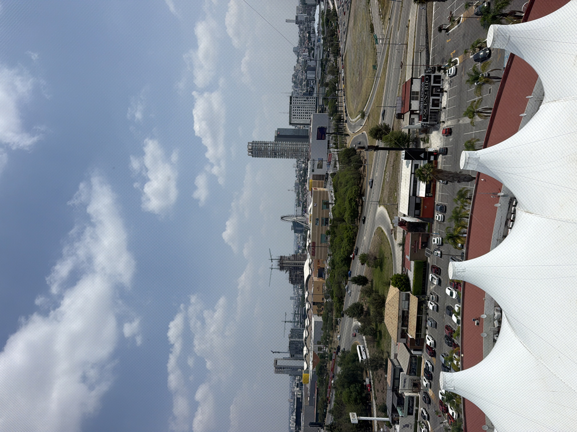 This is an exterior, daytime, medium-angle shot of San Bernardino Tlaxcalancingo, Mexico. The sky is visible with white clouds. The top right corner shows a white, tent-like structure. Adjacent is a view of city streets, parking lots, stores, trees, and high-rise buildings under construction.