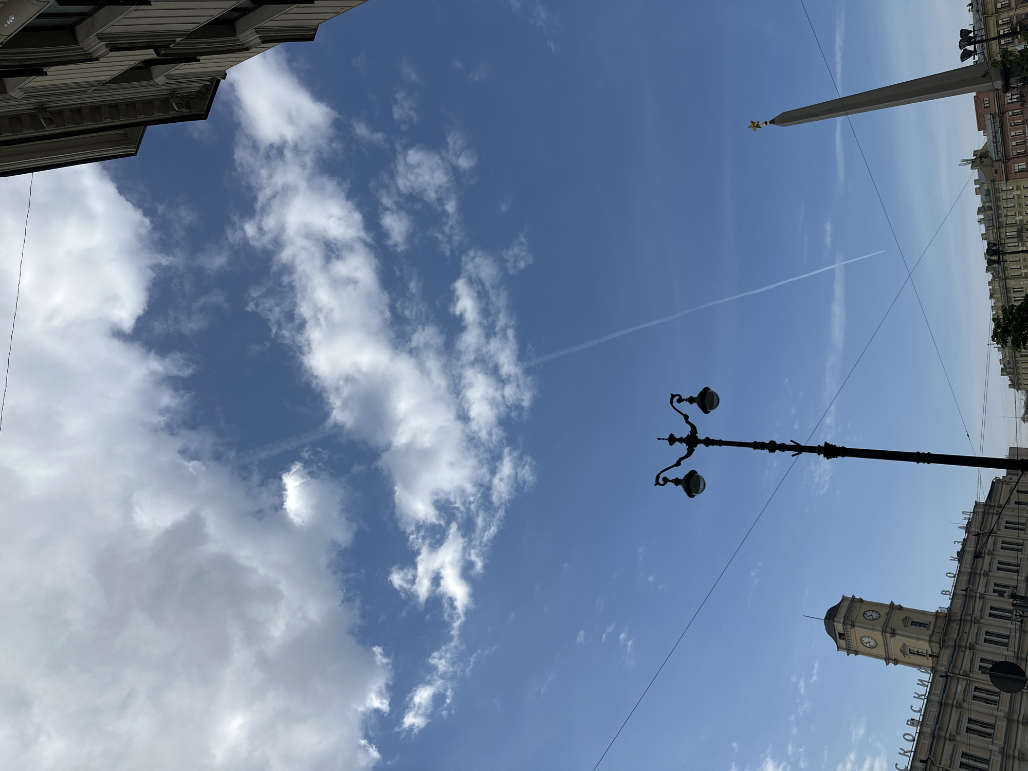 The image shows a view looking upwards in Saint Petersburg, Russia. The sky is mostly blue with scattered cumulus clouds. A contrail from an aircraft stretches across the upper part of the frame. The Alexander Column stands tall in the background. An ornate, black street lamp occupies the center right. The tower of the Vitebsky railway station and the station's sign in Russian ("Витебский вокзал") are on the bottom right. The facades of buildings are partially visible at the top and the right of the image. The scene is devoid of people.