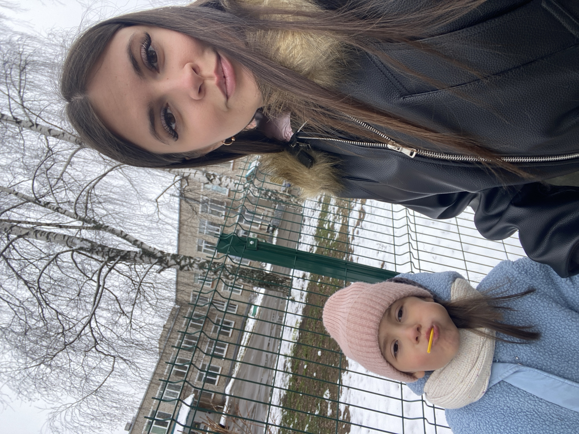 The image shows two individuals outdoors during winter. The taller person, likely a young woman, is positioned in the upper part of the frame, wearing a black leather jacket with fur trim. The younger person, a child wearing a pink knit hat and a light blue jacket, is positioned below, with a yellow lollipop in their mouth. They are standing near a metal fence. Behind the fence, a multistory building can be seen. Snow covers the ground, and a bare tree is visible in the background. The image was taken in the city of Chaikovsky, Russia.