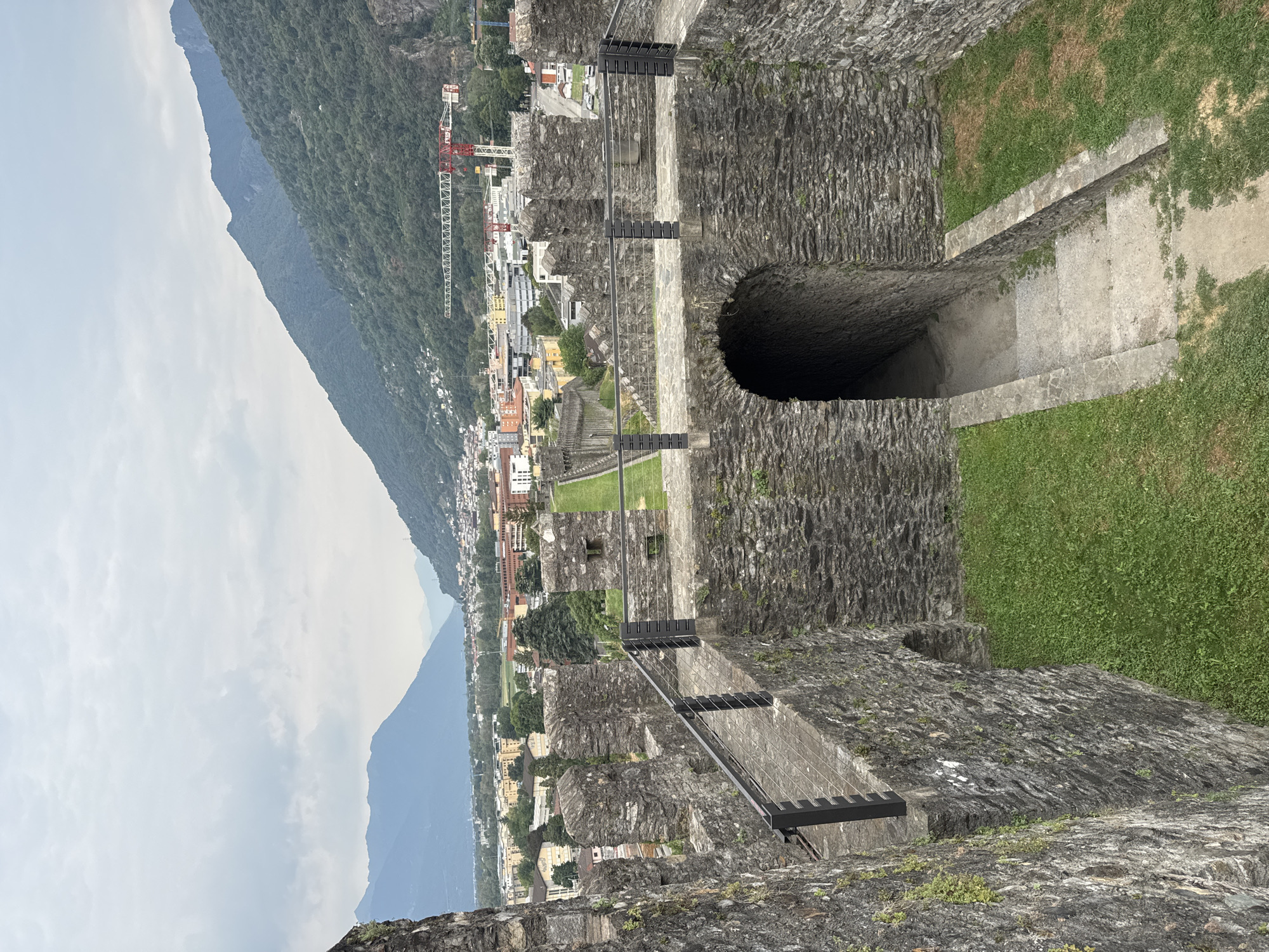 The image shows a view from the battlements of Castel Grande in Bellinzona, Switzerland. The stone walls of the castle are visible in the foreground, with crenellations and embrasures along the top. A pathway with a metal railing runs along the wall, leading to an arched entranceway with stone steps descending into darkness. Beyond the castle walls, the city of Bellinzona spreads out with buildings of various colors and heights. In the background, lush green mountains rise steeply, partially obscured by a cloudy sky. A construction crane is visible among the buildings. The scene is one of historical architecture set against a natural landscape.