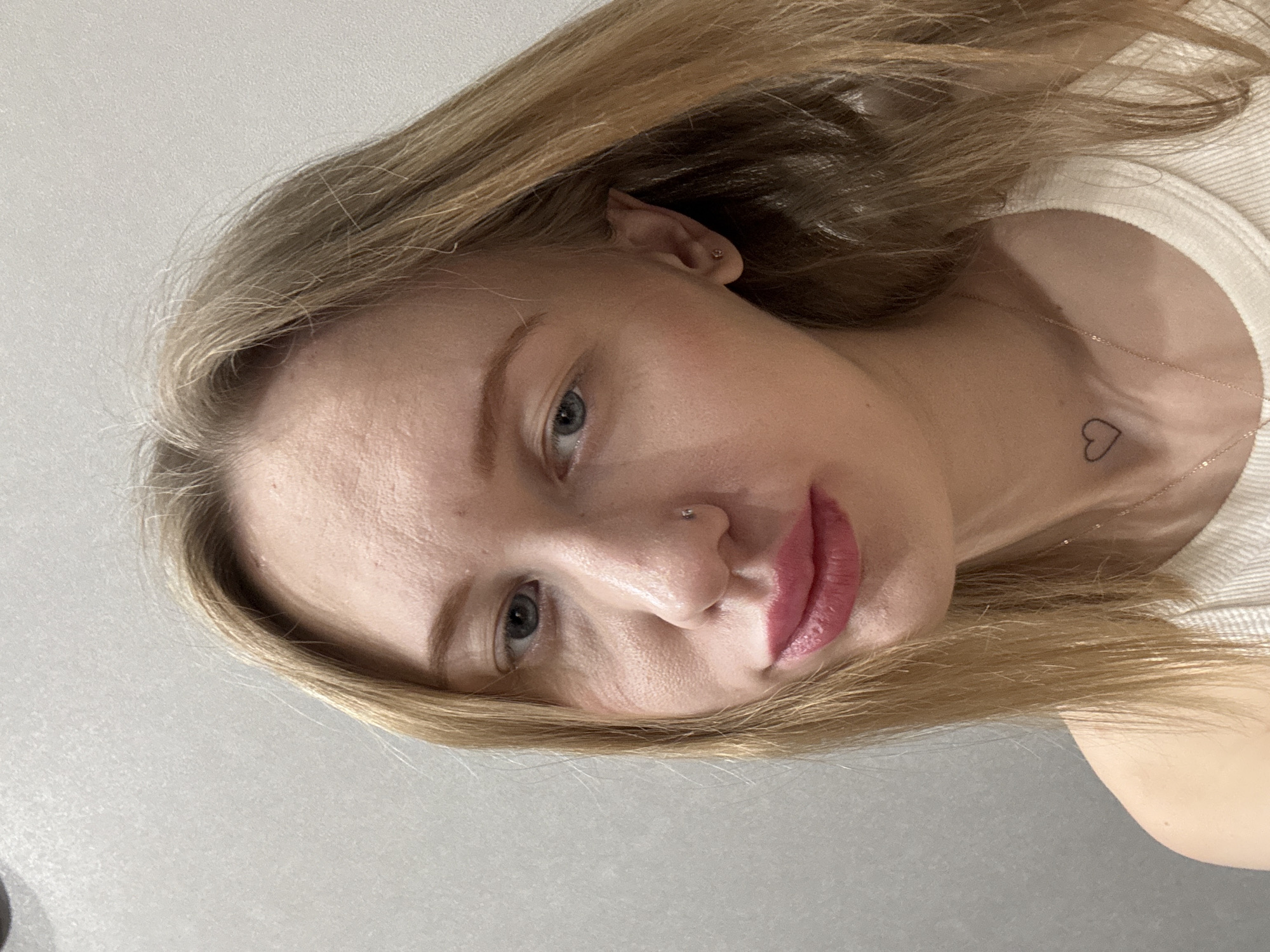 The image presents a close-up portrait of an adult female, situated indoors within the city of Rostov-on-Don, Russia. The subject has light brown hair, blue eyes, and a light skin tone. Her left nostril features a small silver stud piercing. A small, black outline heart tattoo is visible on the lower left side of her neck, above a thin, gold-colored chain necklace. She is dressed in a light-colored, ribbed garment, possibly a tank top or camisole. Her lips are covered in pink lipstick and are slightly parted. The background is a plain, light gray textured wall. The individual is looking directly at the camera.