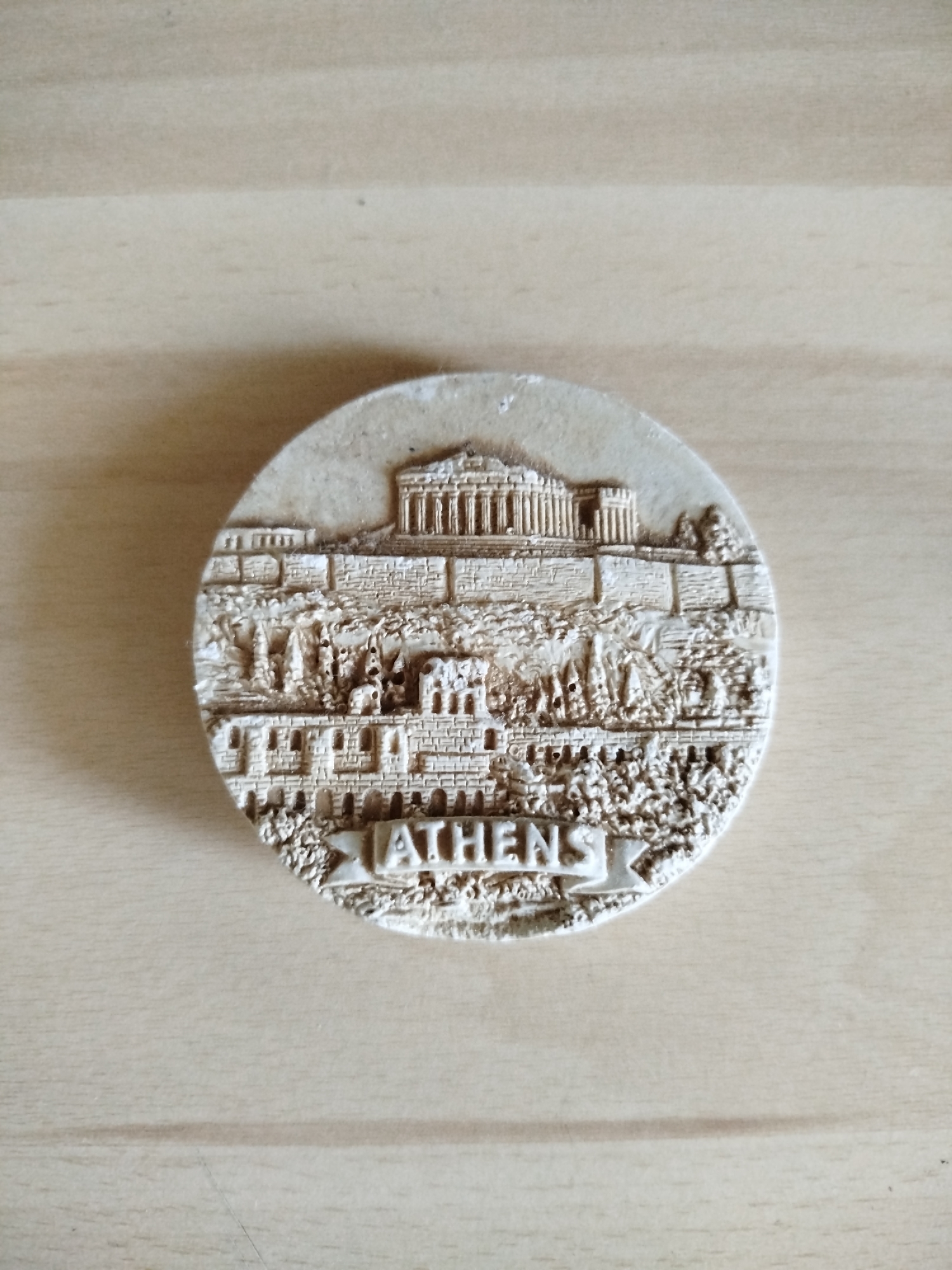 A circular souvenir of Athens, Greece, is centered on a light brown wooden surface. The souvenir depicts the Acropolis and the Parthenon, with the word "ATHENS" prominently displayed at the bottom. There are no people visible. The location is not in Varna, Bulgaria, but Athens, Greece.