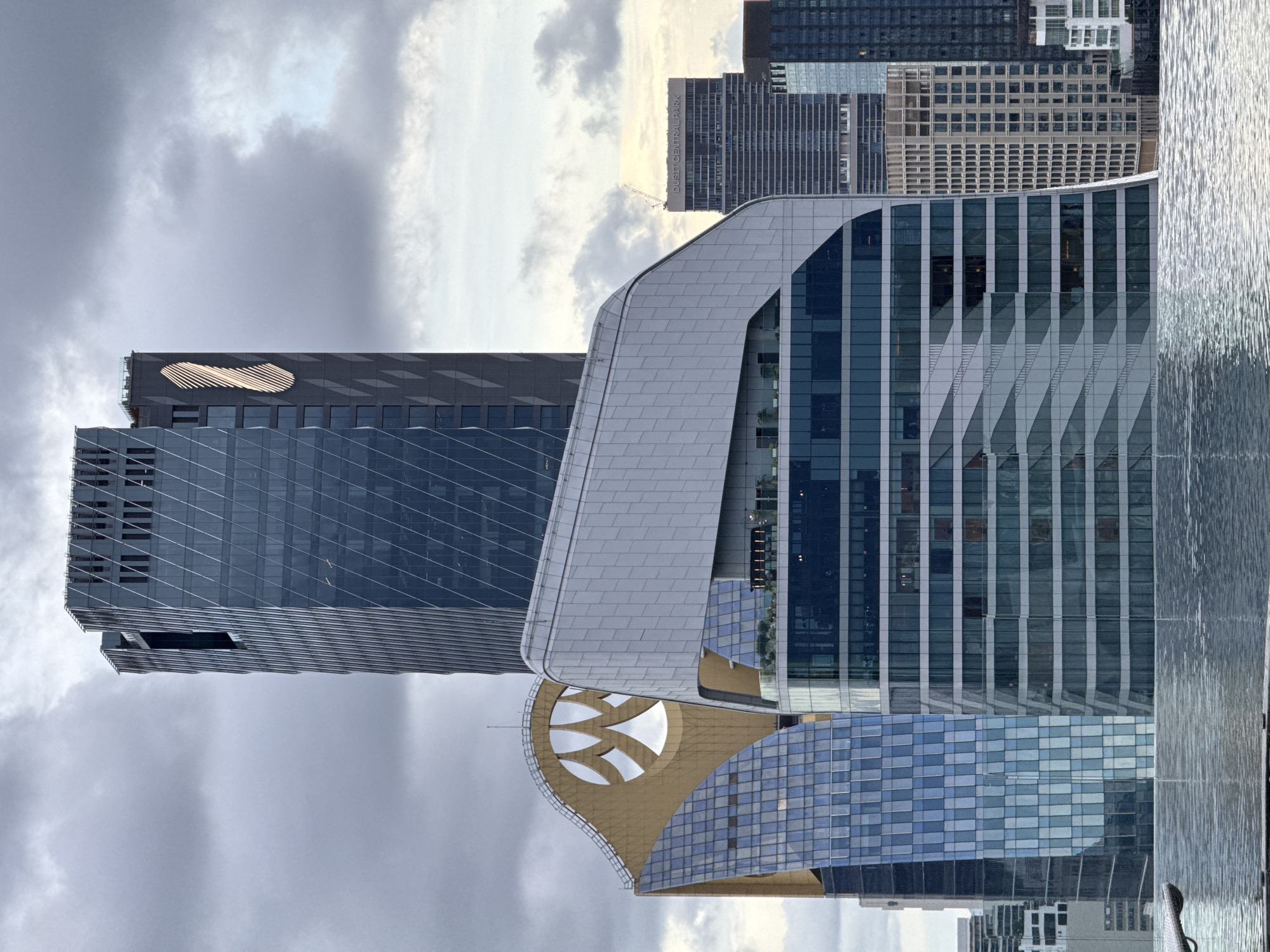 This is an aerial, vertically oriented photograph of a modern cityscape in Bangkok, Thailand, under a cloudy sky. The scene is dominated by contemporary skyscrapers with striking architectural designs.
In the foreground, a curved building with a metallic silver facade features a pattern of geometric shapes. Adjacent to it is a taller building clad in glass, reflecting the sky and surrounding structures. Further back, a dark grey skyscraper with a distinctive logo, a stylized golden oblong with horizontal lines, stands prominently. Behind these, a collection of other high-rise buildings forms the urban backdrop. One building in the middle distance is partially obscured by clouds and has text on it that reads "DUSIT CENTRAL PARK".
The weather appears overcast, with a diffused light suggesting it is either early morning or late afternoon. The buildings' reflective surfaces hint at the tropical climate of Bangkok. The overall impression is one of urban development and architectural innovation. There are no people visible in the frame.
