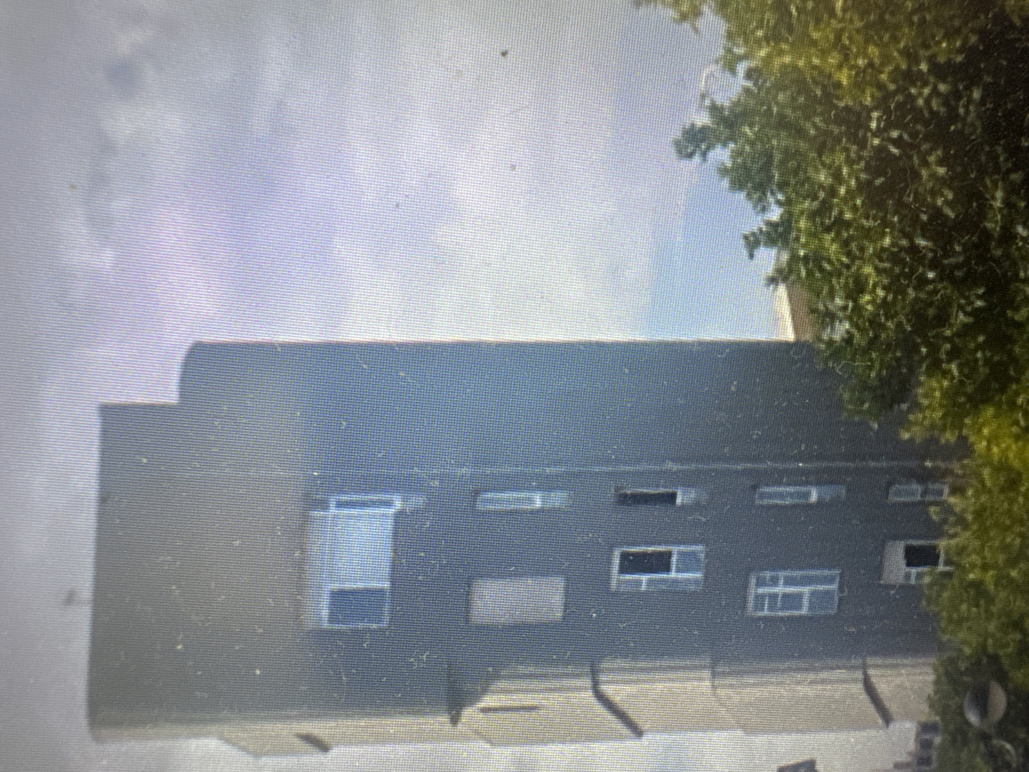 A full shot shows a modern, dark grey, multi-story building with a stepped facade. The building has several windows, some rectangular and others square, with white frames. Some windows appear to have blinds or curtains. The building is partially obscured by lush green trees in the foreground, with their leaves appearing dense and vibrant. In the background, the sky is a pale blue, with some scattered clouds. The building's architecture suggests an urban setting. This building is located in Murcia, Spain, and is the building at Plaza de Europa, number 14.