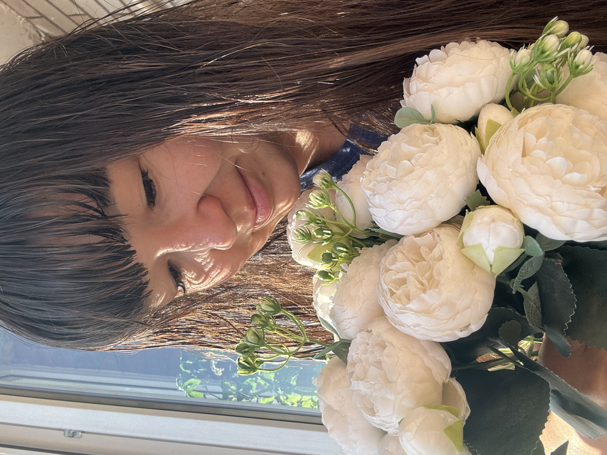 A close-up shot captures a woman holding a bouquet of white peonies and green buds. Sunlight streams in from a window, creating a dappled pattern across her face and dark, wet hair. The woman is looking down at the flowers with a gentle expression. She wears a dark blue top. The background shows a window with greenery visible outside, suggesting an indoor setting, possibly a home, in the city of Chaikovsky, Russia. The overall mood is serene and natural, enhanced by the soft lighting and focus on the delicate flowers.