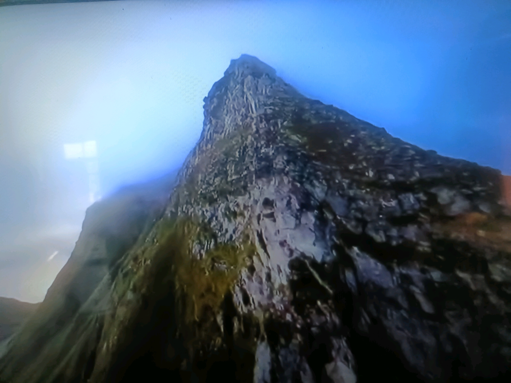 The image shows a jagged, rocky mountain peak reaching towards a bright, hazy blue sky. The mountain face is textured with dark gray rock, interspersed with patches of green and yellow vegetation, suggesting rugged terrain. Some areas of the mountain are obscured by wisps of white mist or clouds, creating a sense of height and atmospheric conditions. The lower left portion of the image reveals more of the mountain's slope, appearing less steep and more covered in vegetation, blending into a bright, washed-out area that could be light or a portion of the sky. There are no people, animals, or man-made structures visible in the image. The scene is natural and dramatic, focusing entirely on the imposing mountain landscape. This scene does not appear to be located in the city of Қасымбек датқа, Kazakhstan, as there are no urban elements or recognizable geographical features associated with that location.