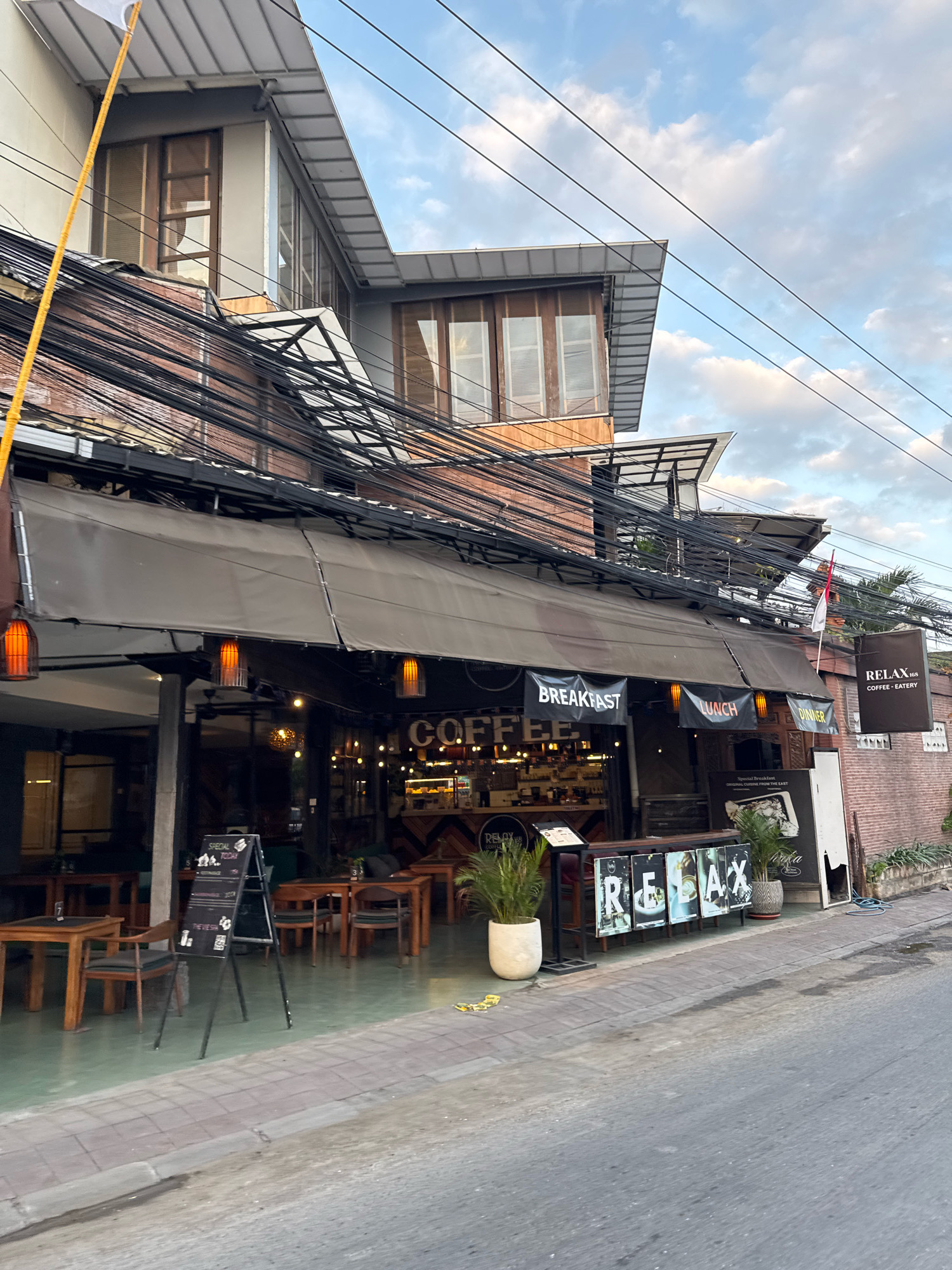 The image shows a street view in Kuta, Indonesia, with a cafe named "RELAX COFFEE - EATERY" prominently featured. The cafe has an outdoor seating area with wooden tables and chairs, and a canopy extending over the entrance. Several illuminated signs are visible, advertising "BREAKFAST", "LUNCH", and "DINNER". Inside the cafe, a counter with food displays and various light fixtures can be seen. A blackboard sign with "SPECIAL TODAY" is placed outside. The cafe's facade is brick and has multiple windows. A flag of Indonesia is visible on the right side of the building. The street is paved with grey stones and asphalt. Overhead, a complex network of black electrical wires stretches across the frame. The sky above is partly cloudy with a blend of blue and white. There are no people visible in the image.