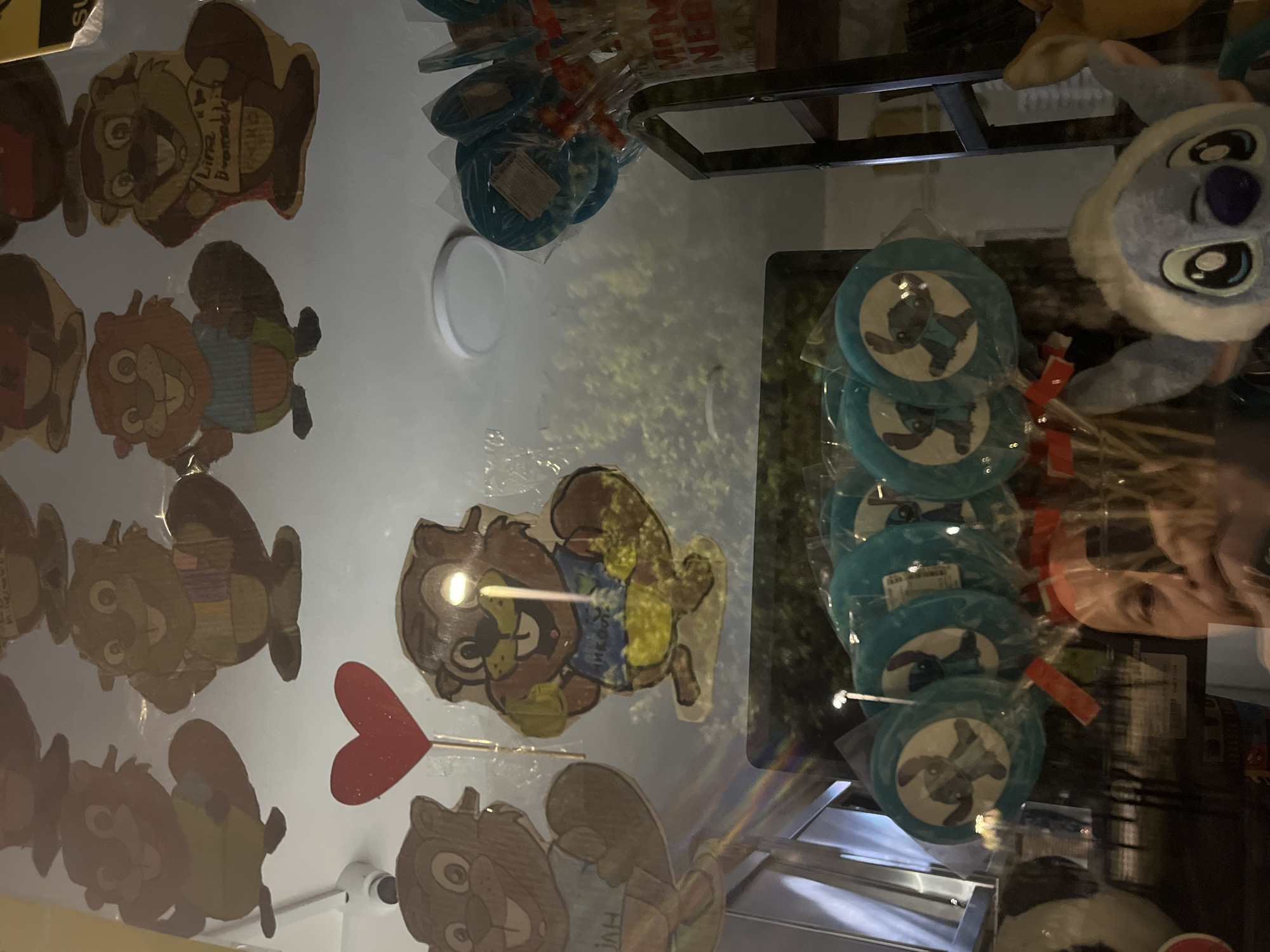 The image shows a display in what appears to be a store or shop in Budva, Montenegro. The display features a selection of souvenirs and merchandise likely targeting tourists. On the left side of the frame, there are several cartoon drawings of beavers or similar animals, possibly hand-drawn or painted on wood or cardboard. Some of these cartoon figures are holding or associated with hearts.
On the right side, there are wrapped lollipops with a cartoon character (possibly Stitch from Disney's Lilo & Stitch) printed on them. The lollipops are arranged in a stack. Adjacent to the lollipops is a plush toy that resembles Stitch.
A person's face is reflected in the glass surface of the display, distorting the view. The background shows a glimpse of the store's interior, including shelves and other merchandise. The lighting in the image is somewhat dim and uneven.