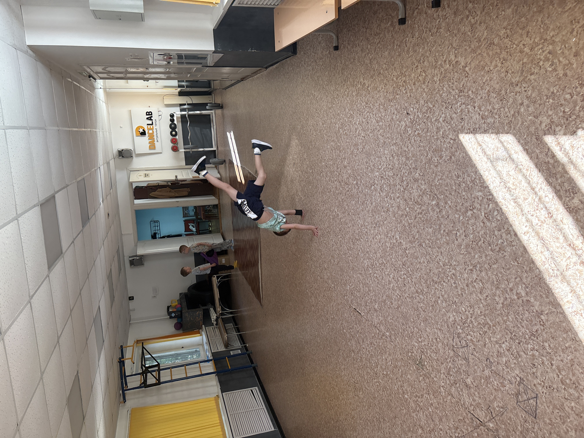 This image captures an interior scene within a multi-purpose room, likely a dance or gymnastics studio, located in the city of Chaikovsky, Russia. The room features light-colored walls, a dark grey wainscoting along the bottom, and a ceiling composed of white acoustic tiles with recessed rectangular fluorescent lighting fixtures. The floor is covered in a speckled, light brown linoleum or vinyl material, showing visible seams and some faint chalk-like geometric markings. Yellow vertical blinds cover windows along the left wall, allowing sunlight to stream in and cast distinct rectangular shadows onto the floor.
In the foreground and mid-ground, a young boy, estimated to be between 6 and 10 years old, is actively performing a cartwheel or handstand. He is inverted, with both hands on the floor and his legs extended upwards and outwards, clad in a light green patterned short-sleeved top, dark shorts with "LEVER" printed on the left leg, and black athletic shoes with white soles.
In the background, positioned to the left of the active boy, two other young boys of similar age are standing still and observing. The boy on the left wears a light-colored, possibly patterned t-shirt and dark trousers. The boy next to him wears a light-colored, patterned t-shirt and grey trousers. They appear to be focused on the boy performing the maneuver.
The room is furnished with various items. Along the left wall, near the observing boys, a blue and yellow gymnastic wall bar system (Swedish ladder) is installed. Below the window, white slatted radiator covers are visible, accompanied by two light-colored wooden benches with metal frames. Adjacent to these, a dark, low-lying object, possibly a large tire or padded equipment, is present, topped with a small collection of colorful balls.
On the back wall, a prominent white sign with an orange star logo displays "DANCELAB" in black text, with the Russian phrase "актуальные танцы" (actual dances) in smaller orange text underneath. Below this sign, a metal weight rack holds several colored disc weights (red, yellow, black) and a barbell rests on the floor in front of it. Two wall-mounted speakers are visible, one near the DANCELAB sign and another on the left wall. Multiple white doors are scattered throughout the room, one of which on the right features a reflective surface. In the far right foreground, two small, light-colored wooden tables with white metal frames are positioned against a wall with dark wainscoting.