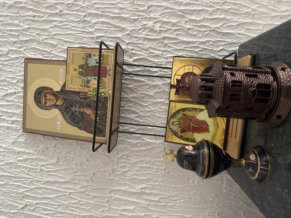 The image displays a collection of religious items arranged on a dark, possibly granite or marble, surface and a two-tiered wire-and-wood shelf unit against a textured white wall, located in Zankovići, Montenegro.
On the upper shelf, a large Orthodox icon of Saint Paraskeva, depicted in dark robes holding a cross, stands upright. Its Cyrillic inscription reads "ПРВ. МАТИ ПАРАСКЕВА." In front of it, a smaller icon portrays an elderly female saint with a staff, situated before a cityscape. A small green and gold Orthodox cross on a silver base is positioned directly in front of the smaller icon.
On the lower surface, to the left, is a dark blue and gold enameled censer with a domed, perforated lid topped by a gold Orthodox cross. It features red and green floral designs. To the right, a dark brown, miniature architectural structure resembling an Orthodox church, complete with a dome and cross, serves as a decorative item or holder. Behind these objects, two more icons are partially visible: one on the left depicts a female saint in red robes with outstretched arms, seated on a cloud against a golden background; the other, largely obscured by the church-like structure, shows a portion of a male saint with a halo and beard against a golden background.