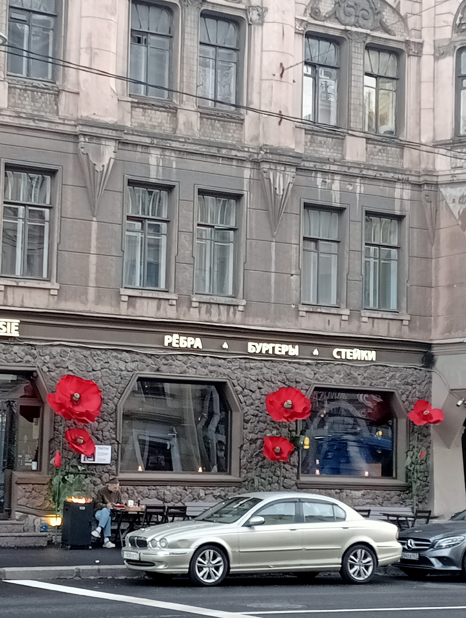 The image shows the facade of a building in Saint Petersburg, Russia, on what appears to be a cloudy day. The building has multiple stories with large windows. The lower level of the building features a stone facade and signage that reads "РЕБРА БУРГЕРЫ СТЕЙКИ," indicating a restaurant specializing in ribs, burgers, and steaks. Red poppy decorations are prominently displayed on the exterior.
In front of the restaurant, several tables and chairs are set up on the sidewalk, with one person seated at a table. A silver sedan is parked in front of the restaurant, and a dark sedan is partially visible to the right. A black heating unit is positioned near the seated individual. The street in front of the building is wet, suggesting recent rain.