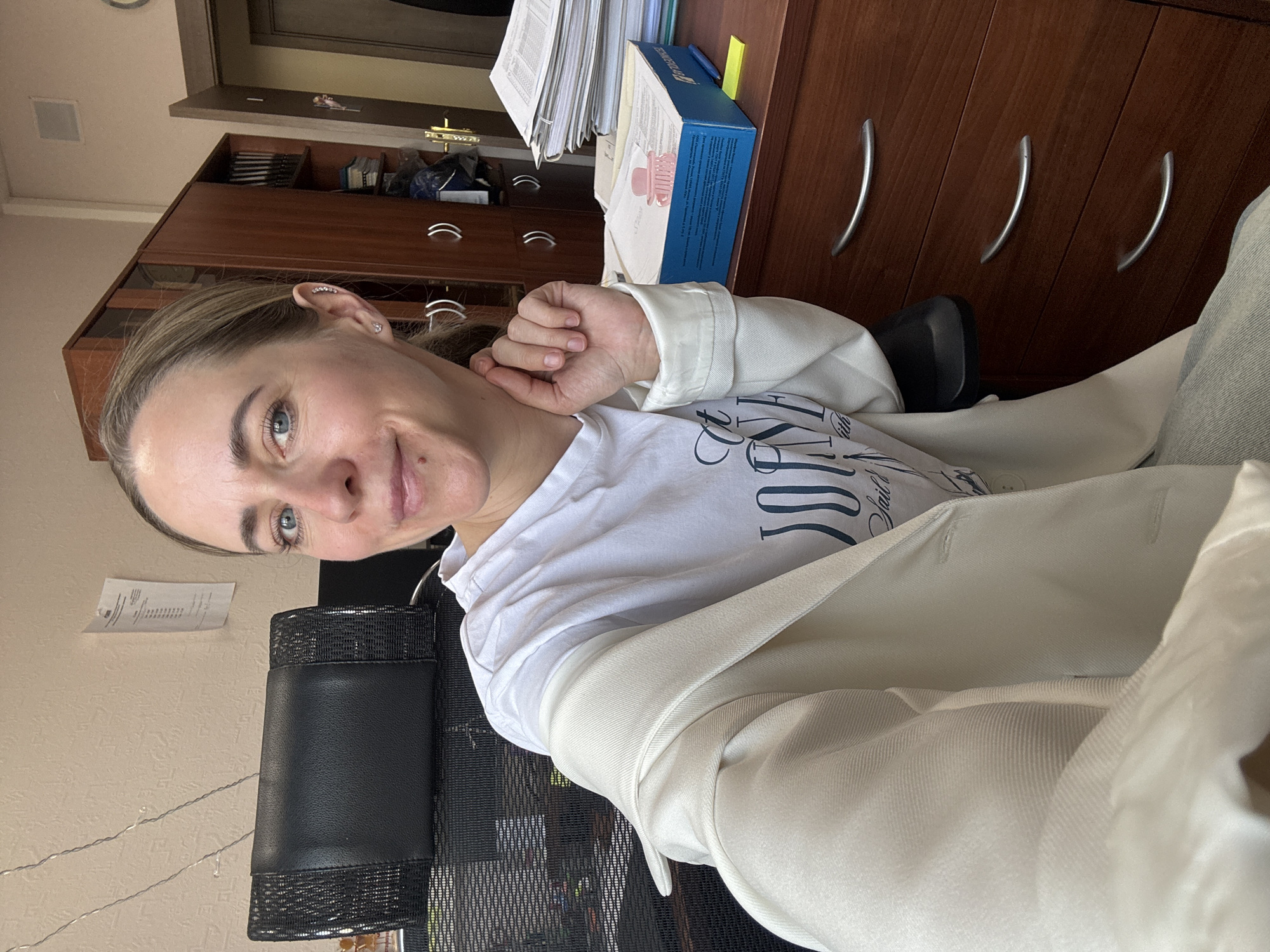 The image shows a woman with light hair and blue eyes, sitting in an office chair. She is wearing a white t-shirt and a light-colored blazer. She is indoors, with a wooden desk and drawers visible behind her, along with stacks of paper and a box. The environment suggests an office space, possibly in Chaikovsky, Russia, although this cannot be definitively confirmed from the image alone. A piece of paper is attached to the wall.