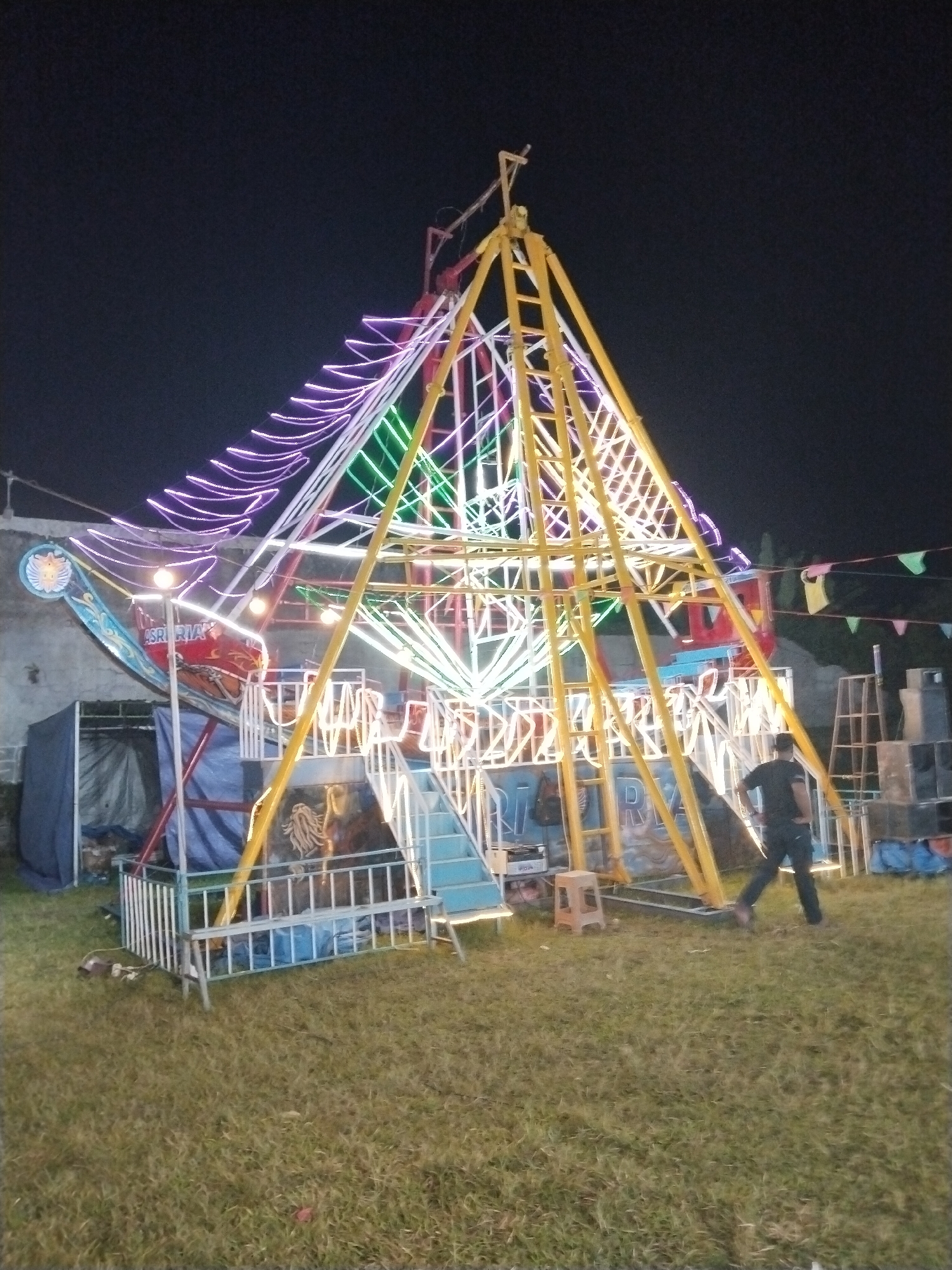 The image shows a small, illuminated Ferris wheel at night. The Ferris wheel is constructed of yellow and white metal beams and is decorated with purple, green, and white lights. It appears to be set up on a grassy field. A man in a black shirt and dark pants is walking near the base of the Ferris wheel. Various objects, including a stool, blue tarps, and stacked equipment, are situated around the Ferris wheel. There is a building visible in the background. The location is Seloromo, Indonesia.