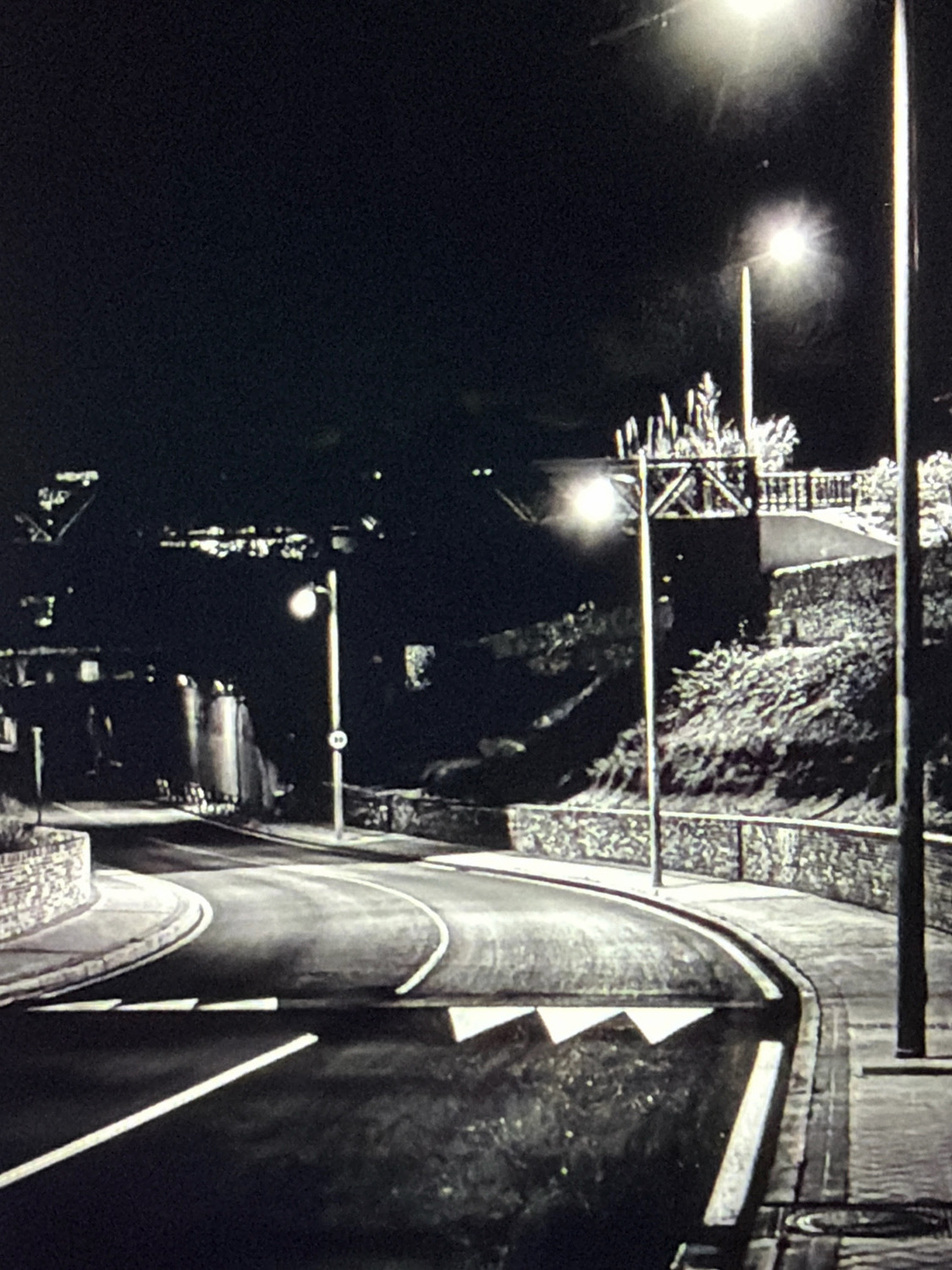 The image depicts a dimly lit, nighttime scene on a winding road. A streetlamp illuminates a section of the asphalt road, which has white markings indicating a pedestrian crossing. The road is bordered by stone walls and foliage on the right, and a curved sidewalk on the left. In the background, structures are dimly visible, including what appears to be an overpass or bridge and industrial-looking cylindrical elements. The overall impression is that of a somewhat isolated urban or semi-urban area. There are no people or animals visible in the frame. The location of Kafr Yasif, Israel, is not discernible from the visual information provided in the image itself.