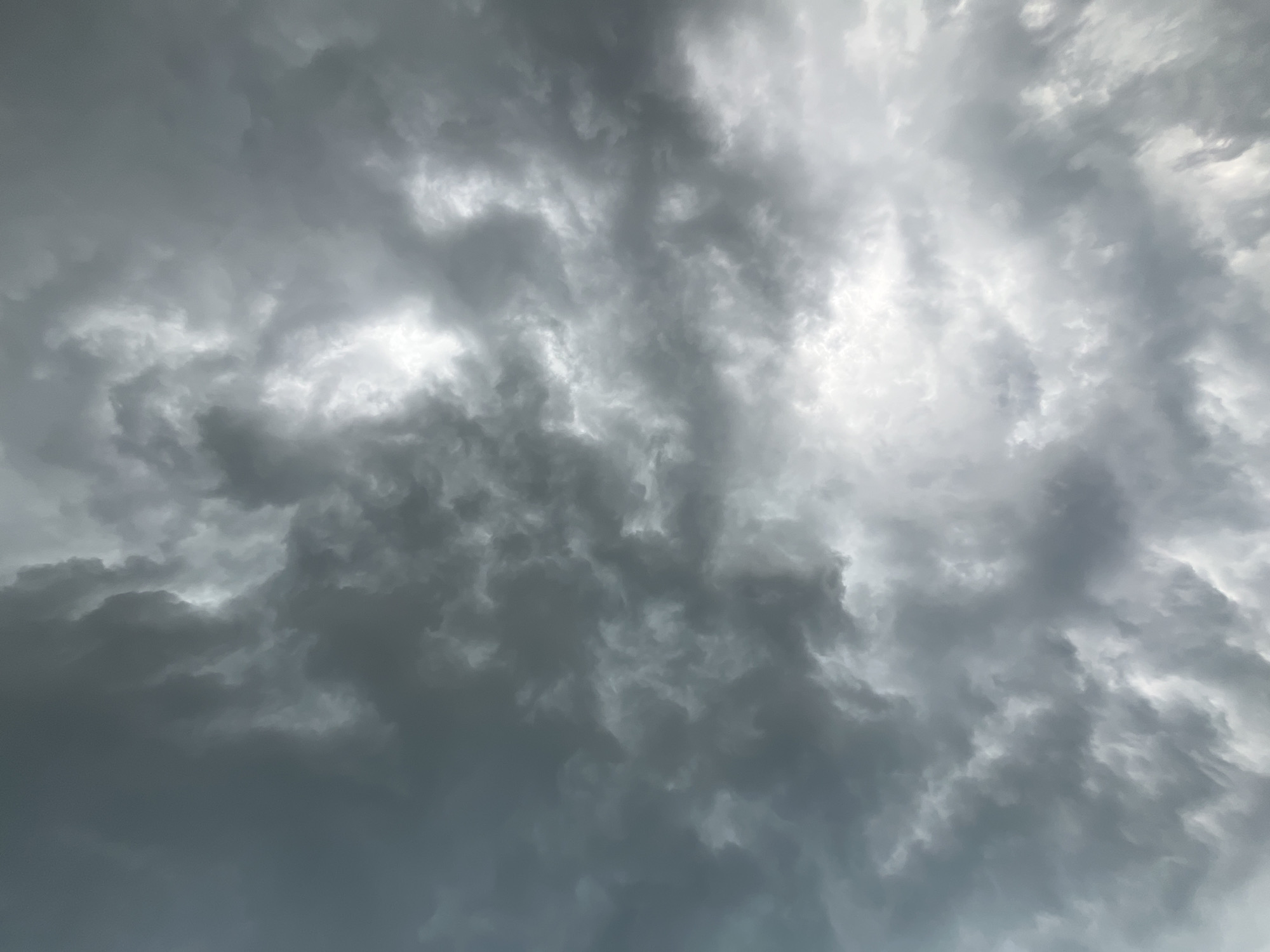 The image shows a sky filled with dark, dramatic storm clouds. There are bright patches where the sunlight is breaking through, creating a contrast with the darker, more ominous clouds. The clouds are dense and appear to be moving, with swirling patterns and varying textures. The overall impression is one of an impending storm. There are no people, subjects, or objects visible in the image, nor any specific environmental details to indicate the location in the city of Маяк, Russia.