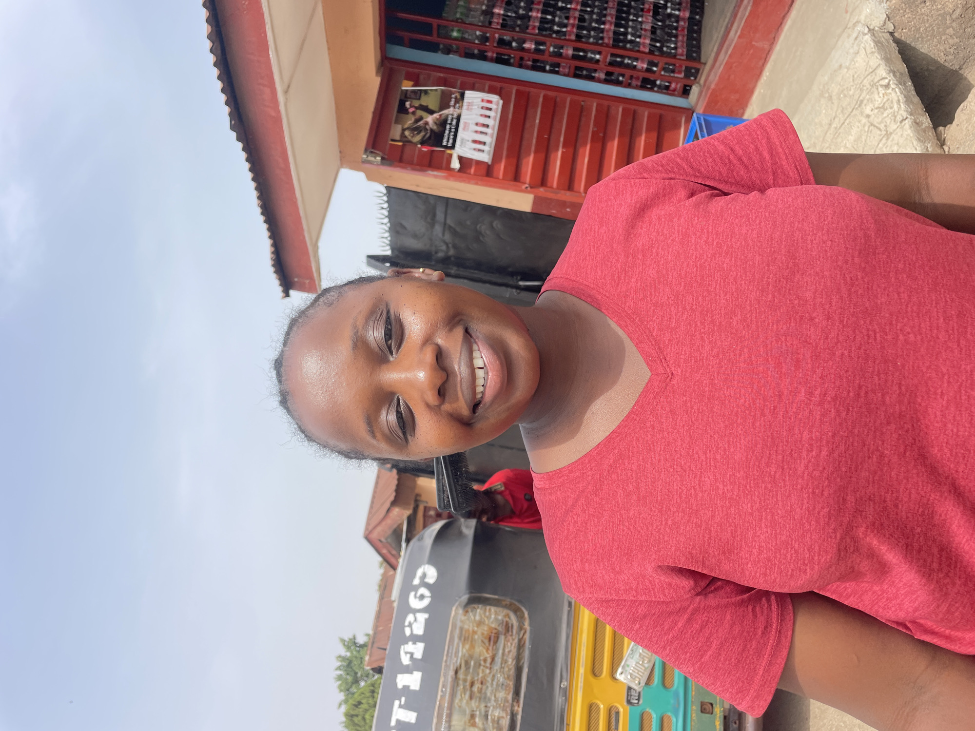A woman with dark skin and hair pulled back smiles at the camera. She is wearing a red t-shirt. Behind her is a commercial building with a red painted front. A three-wheeled vehicle, common in Nigeria, is partially visible. The vehicle's registration number is partially obscured but appears to be T-11263. The sky is light blue. Based on the vehicles and architecture, the location is likely in Jos, Nigeria.