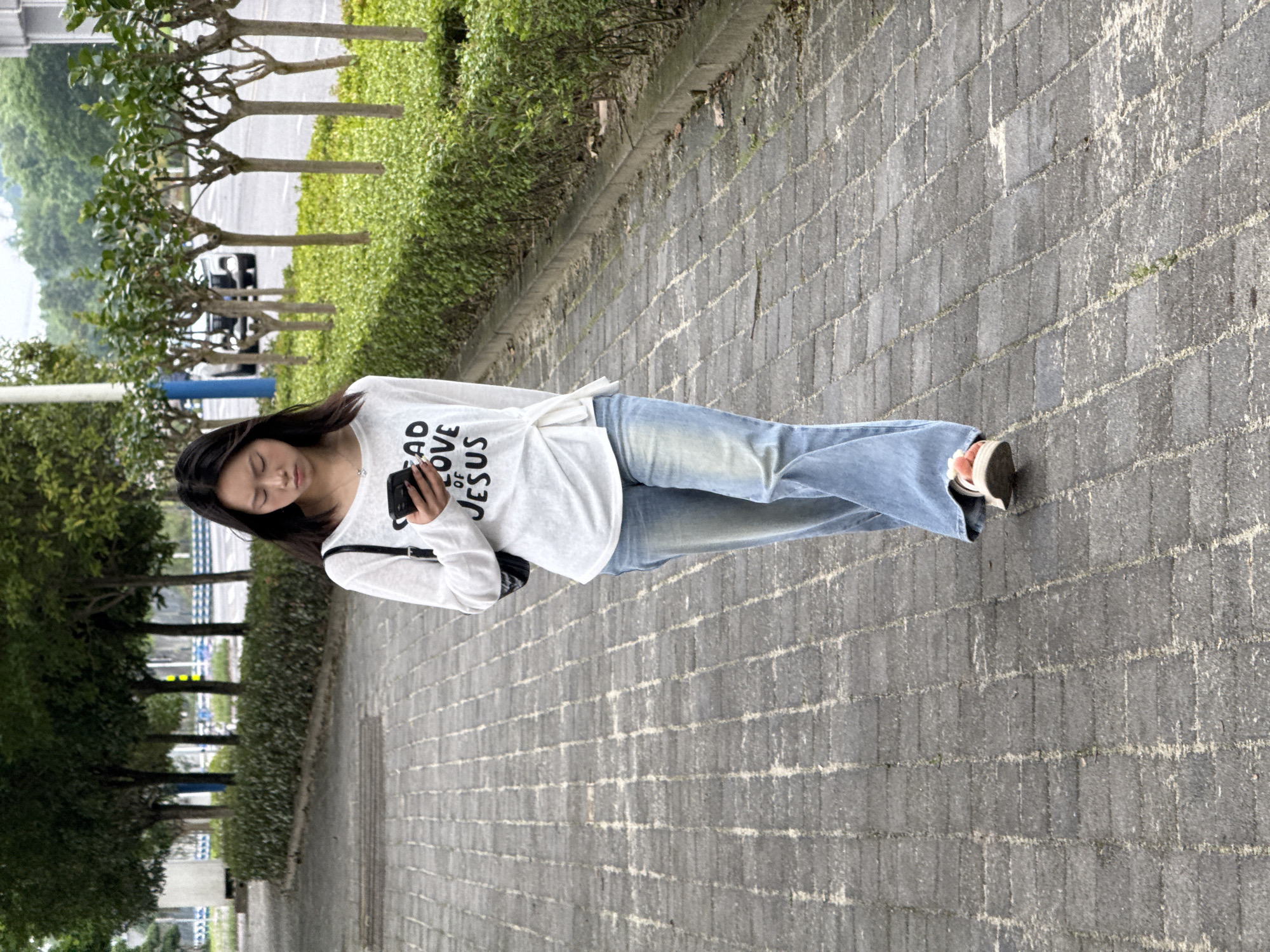 A young adult woman stands on a gray brick-paved sidewalk. She is wearing light-wash blue jeans, a white long-sleeved t-shirt with the printed words "Read of Love of Jesus" and sneakers. She holds a mobile phone in her hand and looks at the screen. A black purse hangs from her shoulder. To her left is a strip of green foliage and sapling trees that recede into the background. In the distance on the left of the frame, there are glimpses of vehicles and urban infrastructure. This image was taken in Jiangjin, China.