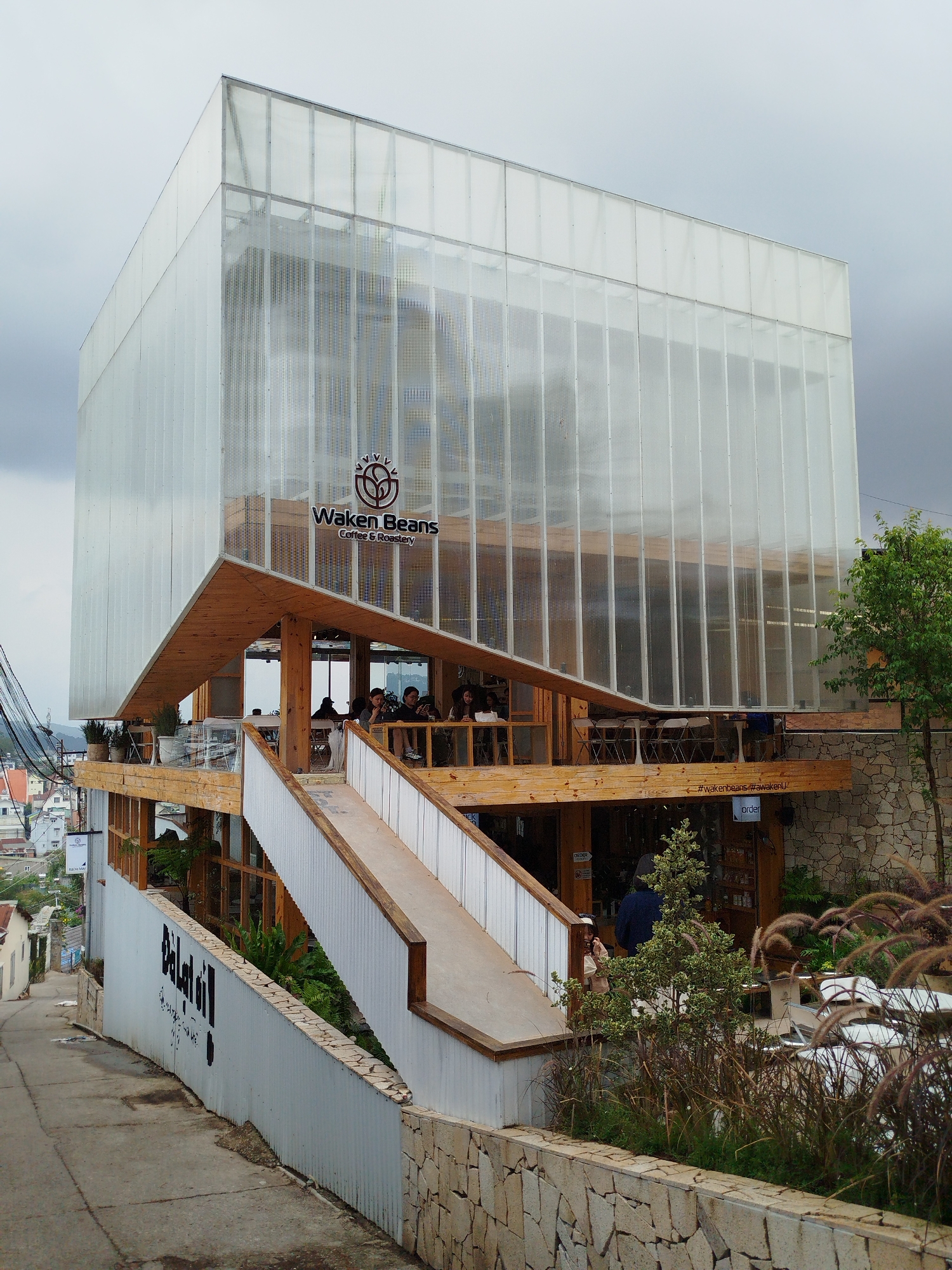 The image displays the Waken Beans Coffee & Roastery, a multi-level cafe nestled into a hillside along a sloped street in Da Lat, Vietnam. The building exhibits a striking contemporary design; its upper two stories are enclosed by translucent, corrugated panels, giving it a bright, ethereal appearance, much like a modern greenhouse. Below this, the structure features open and semi-enclosed wooden terraces, supported by visible timber beams and incorporating stone-clad retaining walls, harmonizing with the natural terrain.
Several patrons are visible on the wooden terraces, seated at tables, suggesting a relaxed atmosphere for socializing and enjoying beverages. A wide, white and wooden ramp or staircase provides access from the sloped concrete road in the foreground to the various cafe levels. Along the roadside, a white corrugated metal wall is prominently marked with large, black painted letters spelling "DaLat!!!", alongside smaller, less legible text.
The scene is captured on an overcast day, indicated by the soft, diffused light and a gray sky. Overhead power lines are visible on the left side of the frame. In the background, numerous houses and buildings sprawl across the hills, characteristic of Da Lat's hilly landscape. Lush greenery, including potted plants on the terraces and ornamental grasses near the base, enhances the natural aesthetic. Key visible text includes the "Waken Beans Coffee & Roastery" logo on the upper facade, and hashtags such as "#wakenbeans #awakenU" and the word "order" inscribed on a wooden beam of a lower terrace.