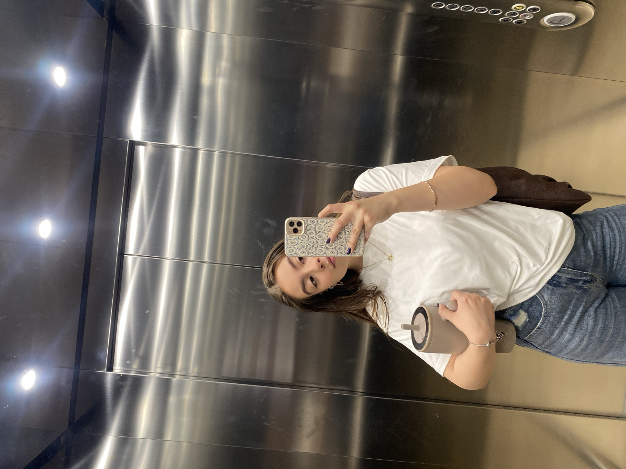 A young woman is depicted taking a mirror selfie inside an elevator. She stands facing forward, with her head slightly tilted and eyes looking directly into her smartphone, which she holds up with her right hand. Her phone has a patterned case. In her left hand, she holds a light-colored tumbler with a straw.
She is wearing a plain white t-shirt and light blue, ripped jeans. A dark brown shoulder bag is slung over her right shoulder, visible behind her. Accessories include a thin gold bracelet on her right wrist, another bracelet on her left wrist, and a delicate necklace with a small pendant. Her fingernails appear to be painted a dark color.
The elevator interior features highly reflective, brushed metallic walls, likely stainless steel, which show prominent reflections of bright artificial overhead lights. The texture of the metal panels is visible, with horizontal lines separating them. In the upper right corner of the image, an elevator control panel is visible, featuring several small, round, dark buttons and a larger, possibly illuminated, button with a downward-pointing arrow.
The overall lighting is bright due to the artificial sources and reflective surfaces. The setting is clearly an indoor elevator. The woman's expression is neutral and posed for the selfie. No specific text is clearly legible on the control panel beyond the shapes of the buttons and the down arrow symbol.
