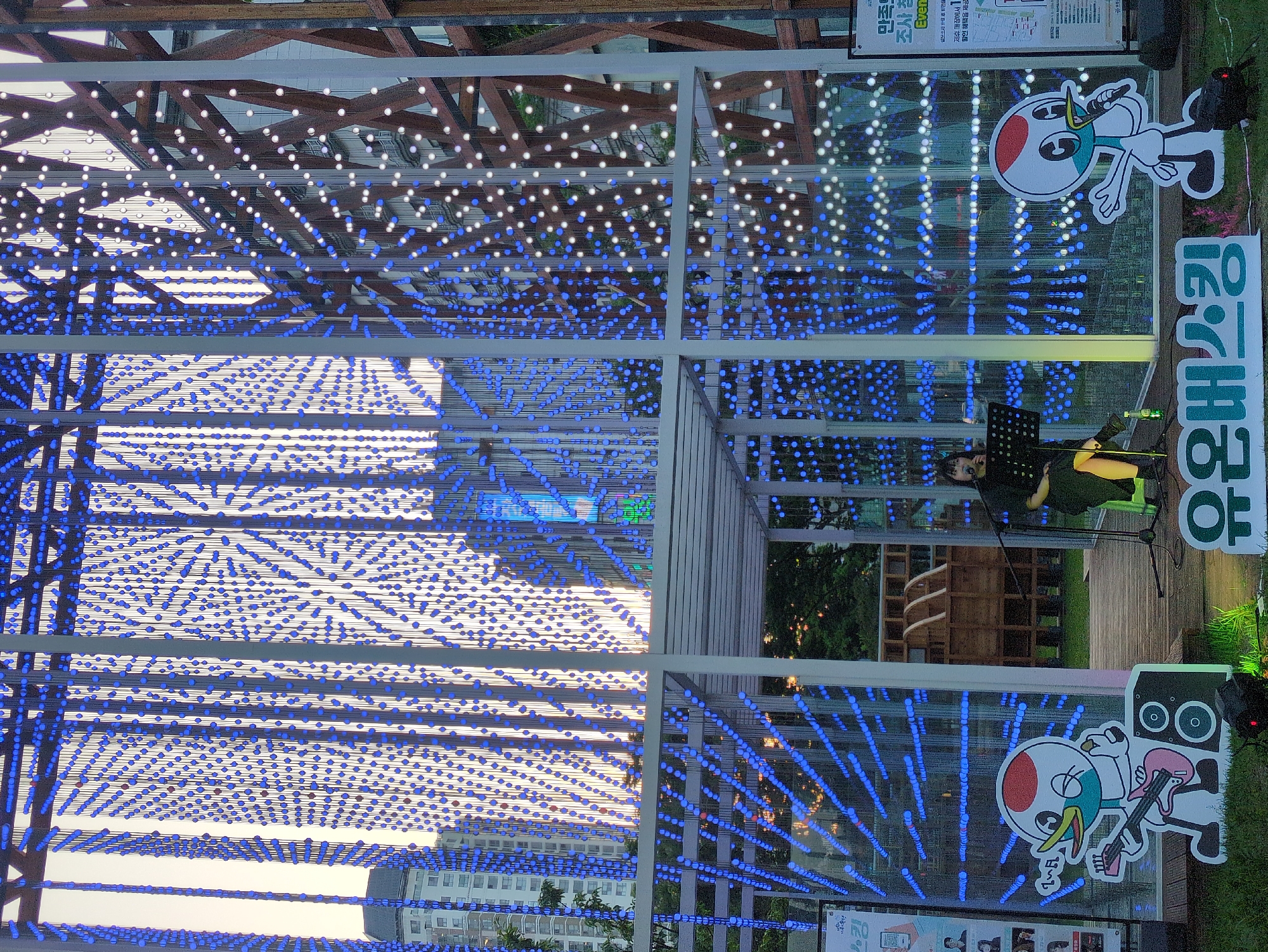 Here's a description of the image:
The image shows a musical performance taking place within an art installation or decorated pavilion, likely at night or during twilight. The structure is adorned with numerous strings of small, illuminated LED lights, mostly blue with some white sections. The strings of lights create a matrix-like or dimensional effect.
A person, presumably a musician, is positioned within the structure with a music stand and what appears to be a keyboard or similar instrument.
The location appears to be in Daejeon, South Korea. Signs/posters with Korean characters are present. There are also cartoon mascots or promotional characters displayed near the structure. In the background, buildings can be seen in the distance, indicating an urban or suburban setting. There is a wooden structure next to the stage.
