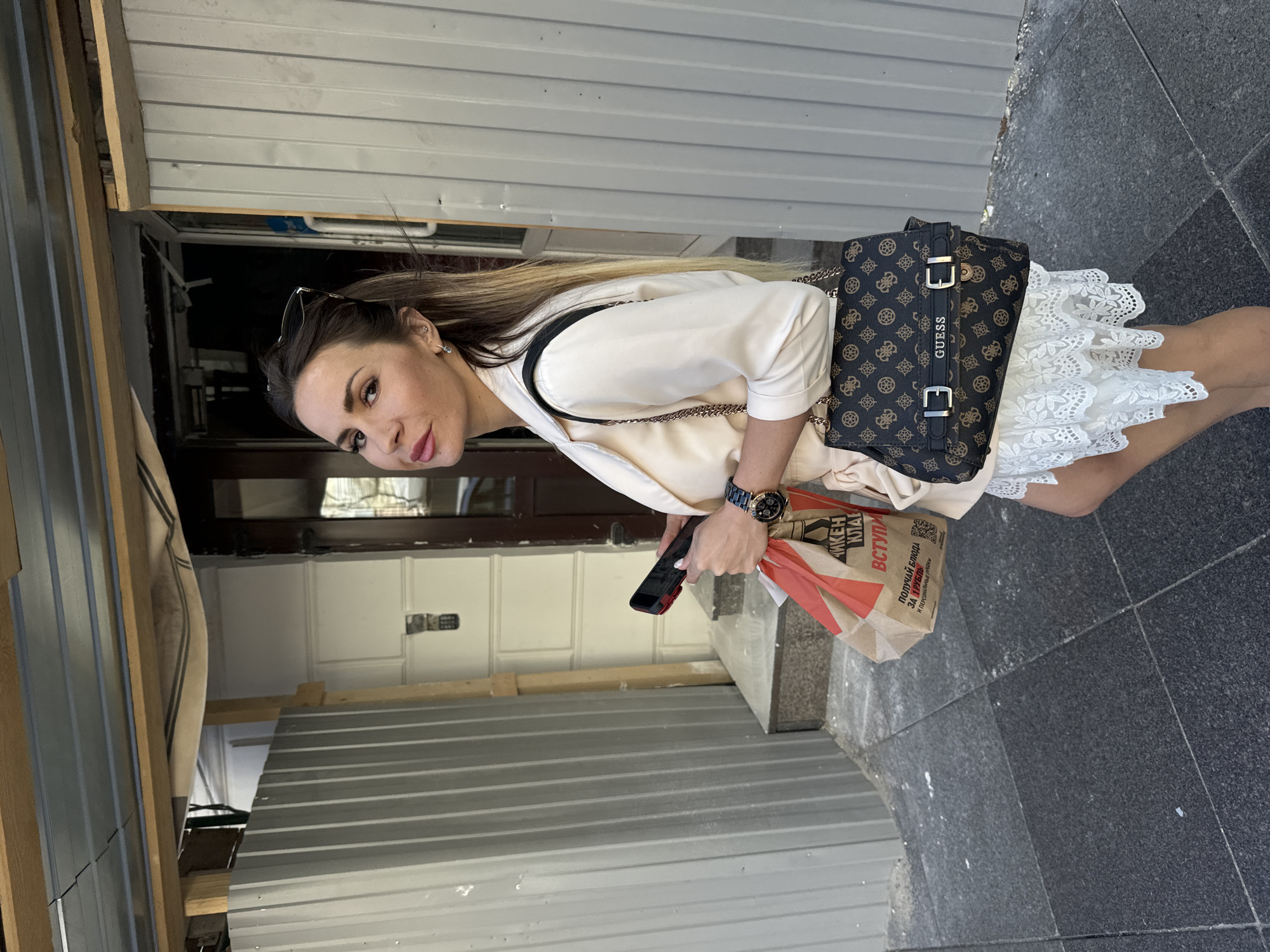 A woman with long blonde hair is standing outside a building. She is wearing a light-colored shirt and a white lace skirt. She is carrying a brown and black patterned "Guess" brand purse and holding a smartphone and a paper bag. The bag has Russian text on it, indicating a food purchase. The building appears to be under construction or renovation, with corrugated metal siding visible. The ground is paved with dark tiles. The lighting suggests it is daytime. The woman is looking at the camera with a slight smile.